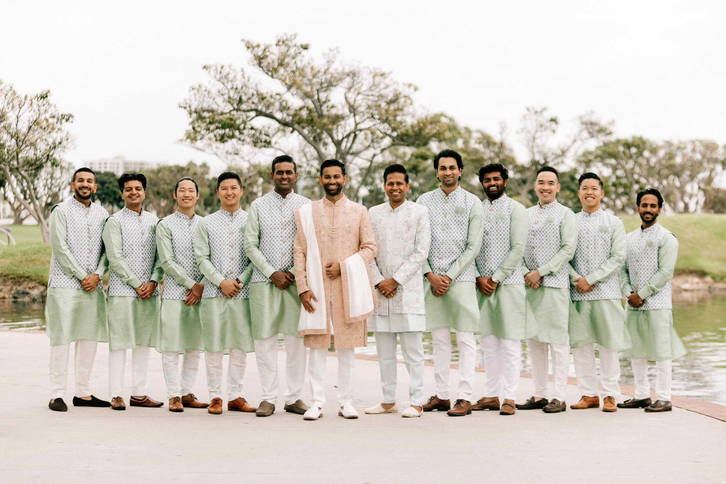 A group of eleven men in traditional Indian attire standing outdoors near a body of water with trees in the background, smiling at the camera.