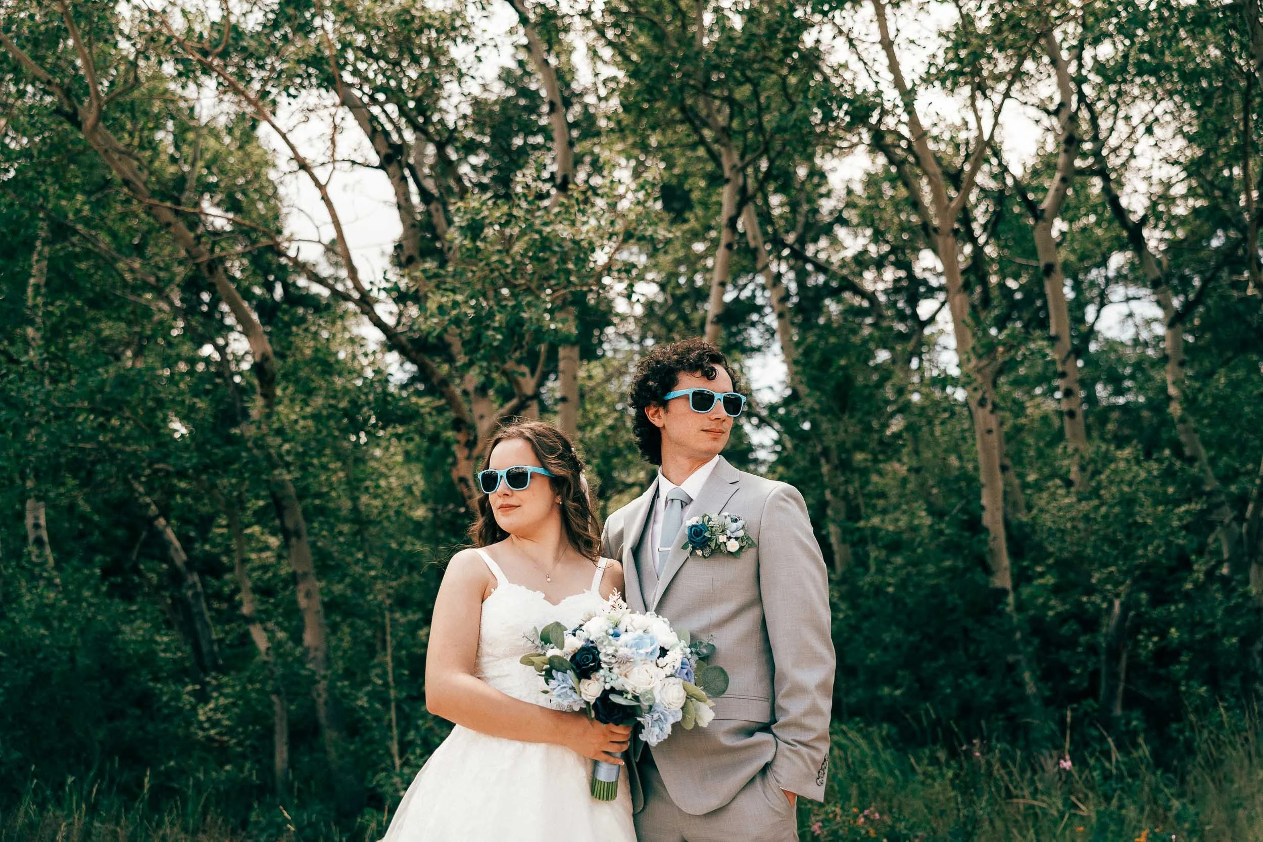 A bride and groom stand outdoors in a wooded area, with the bride holding a bouquet of blue and white flowers, both wearing sunglasses and dressed in wedding attire.