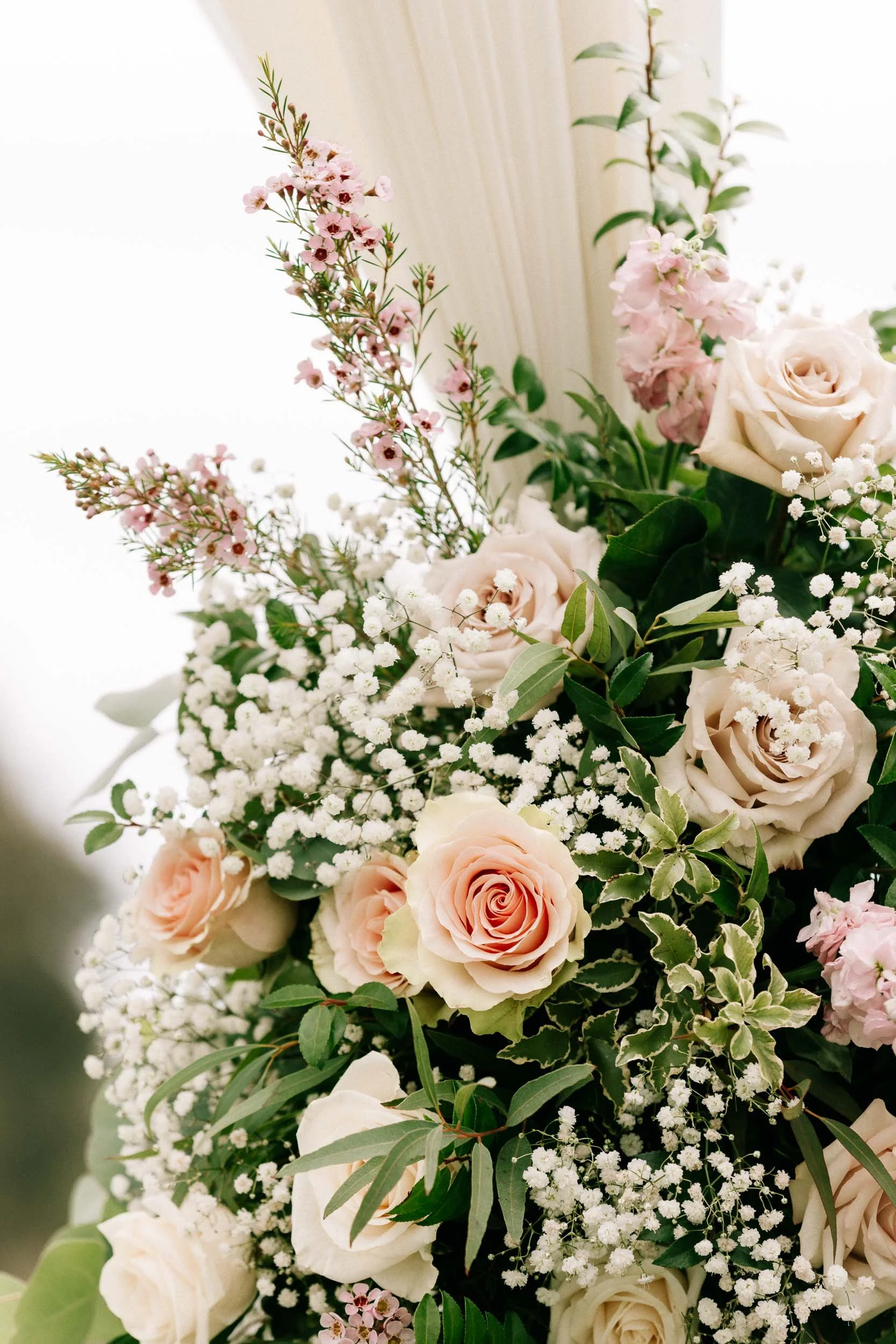 A bouquet of light pink and white roses, pink waxflower blossoms, baby's breath, and various green leaves.