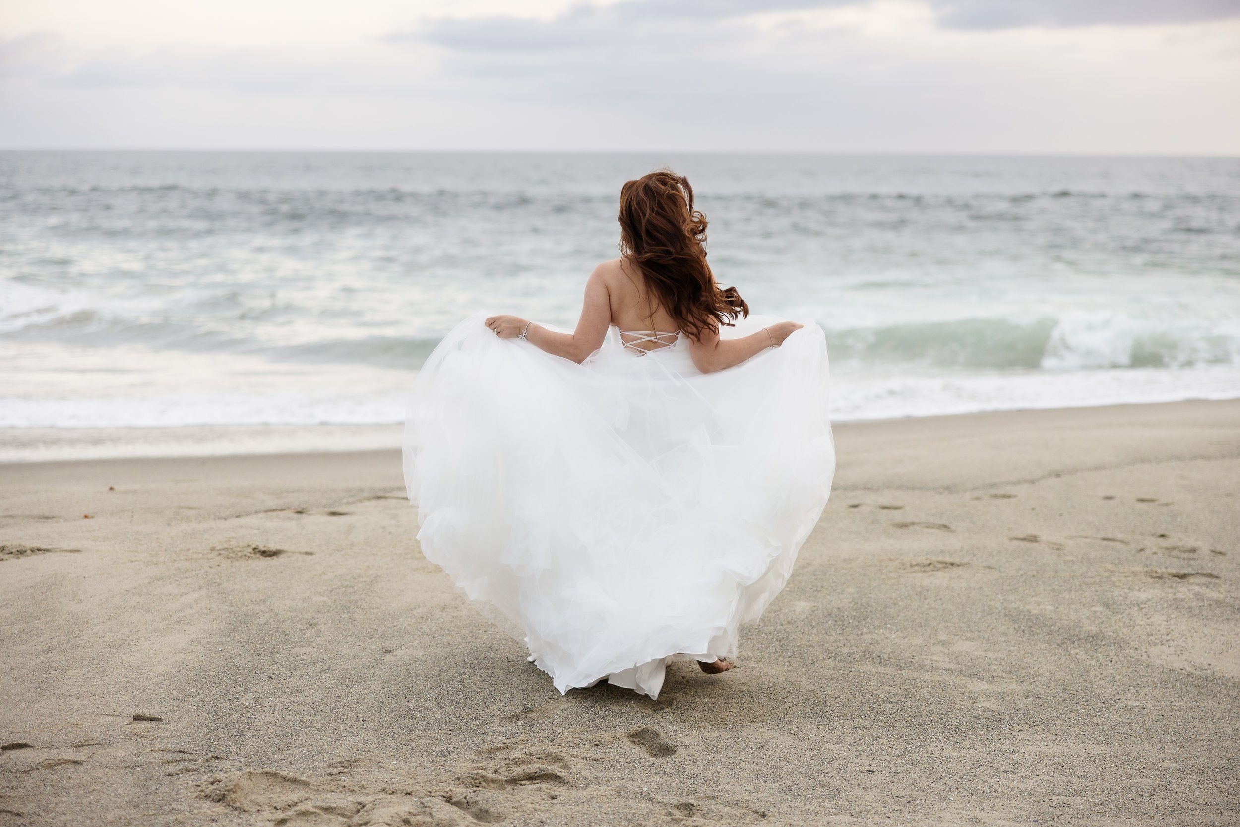 A woman in a white wedding dress walking on the beach with the ocean in the background.