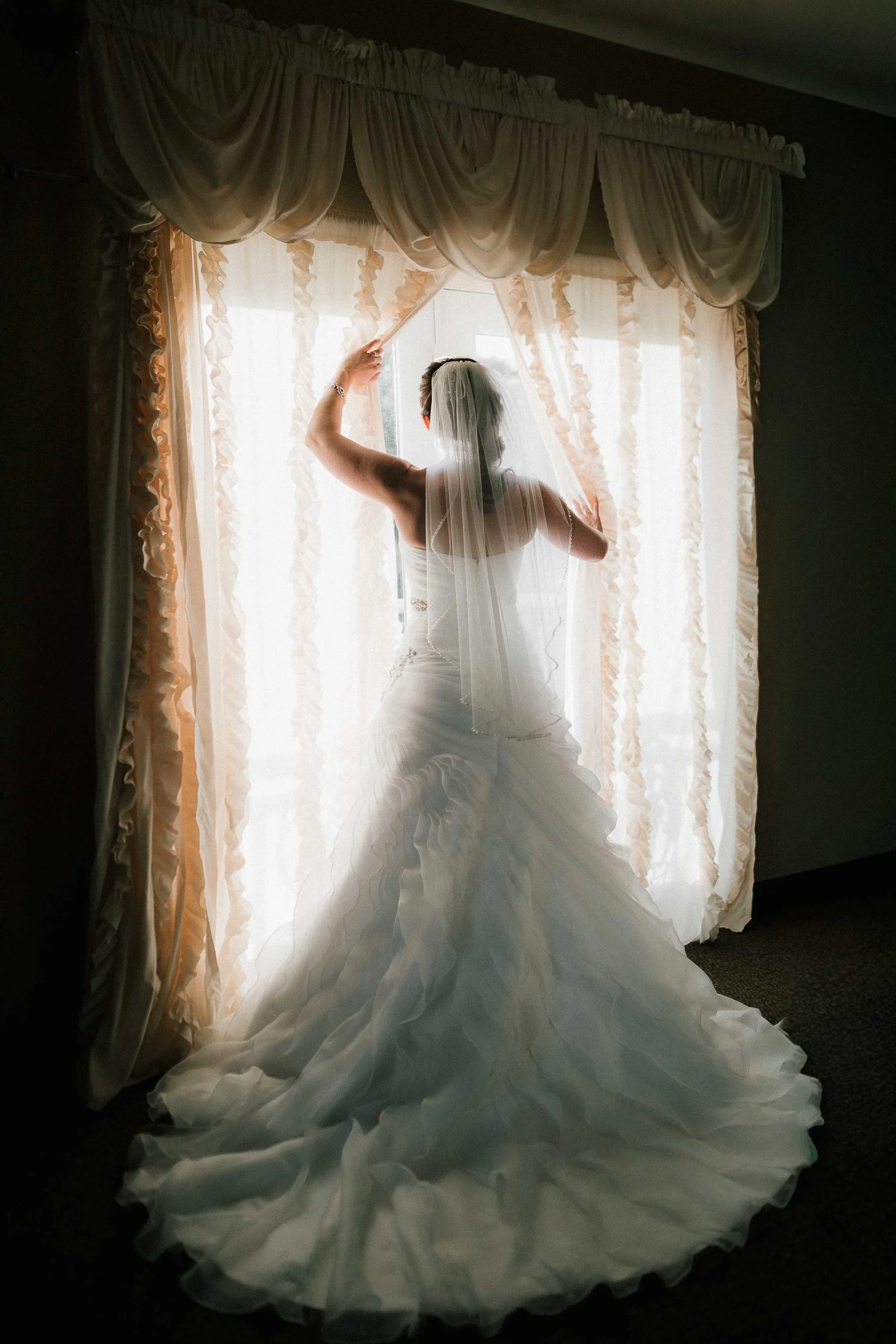 A bride in a wedding dress and veil looking out a window with light curtains, adjusting the curtain.