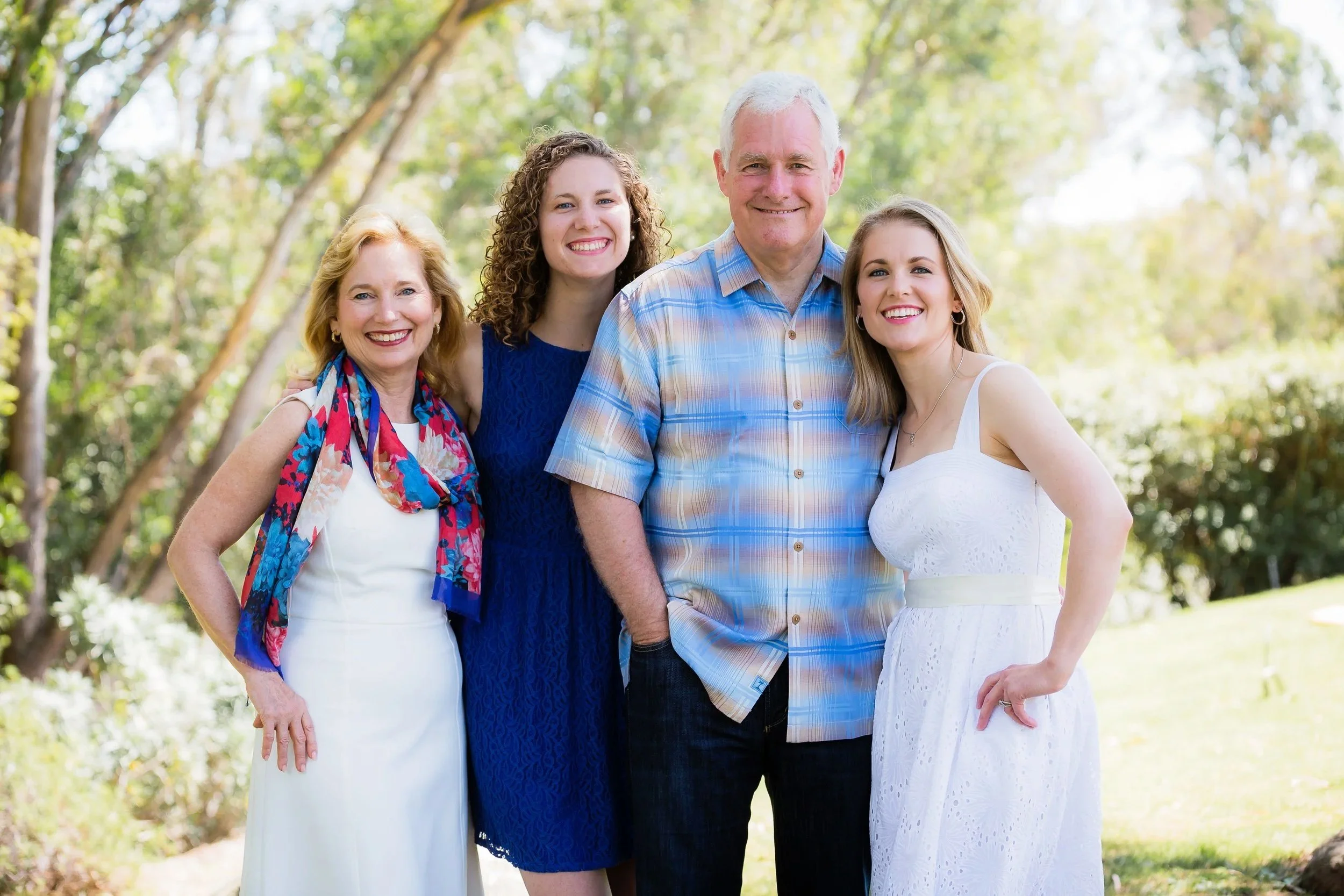 A family of five smiling outdoors in a park with trees in the background.