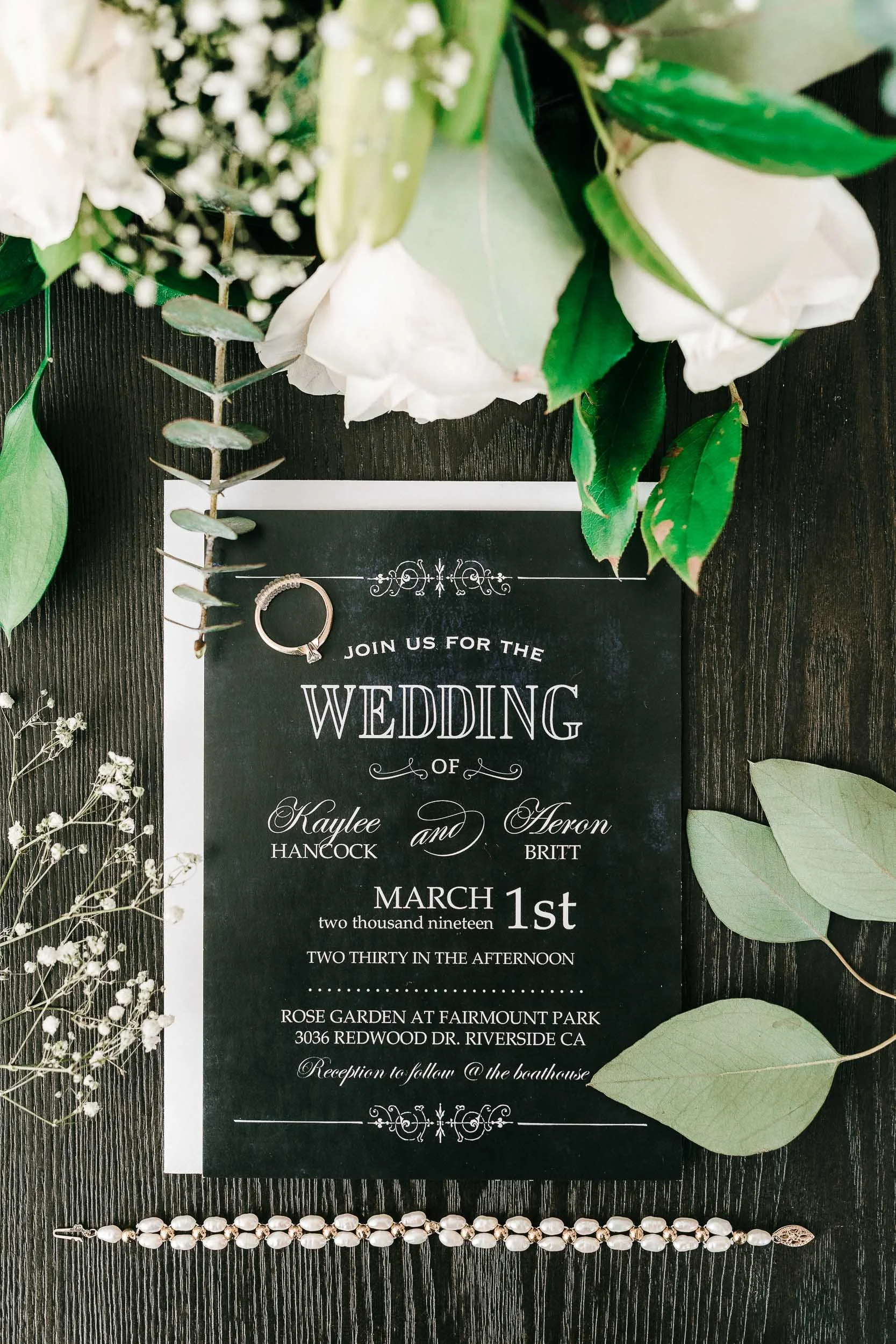 Wedding invitation surrounded by white flowers and green leaves, with a pearl necklace below the invitation on a black wooden surface.