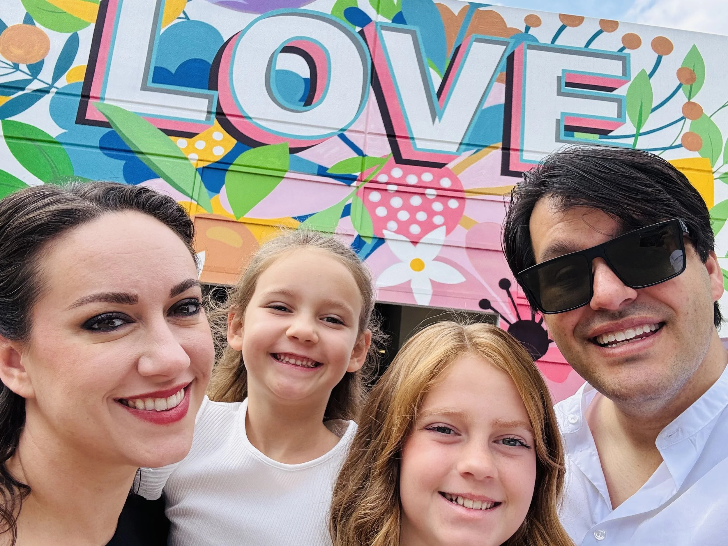 Close-up of a smiling woman with dark hair, a smiling girl with blonde hair, and a man with dark hair and sunglasses, in front of a colorful mural with the word "LOVE" and floral patterns.