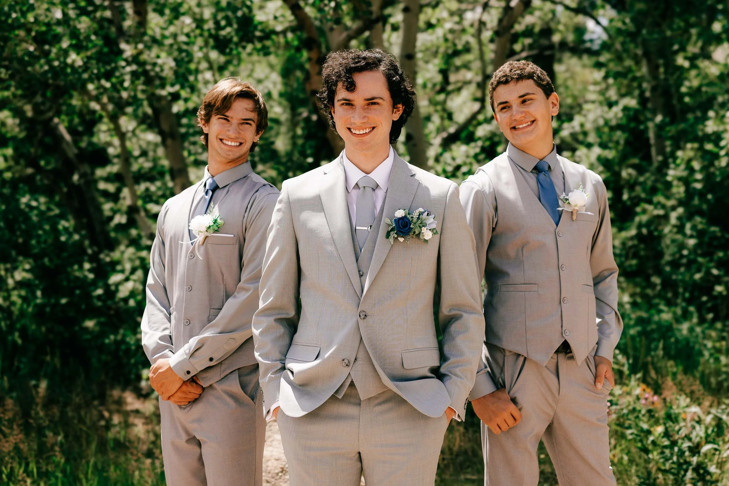 Three young men dressed in formal suits, smiling outdoors in a park with green trees at a wedding.
