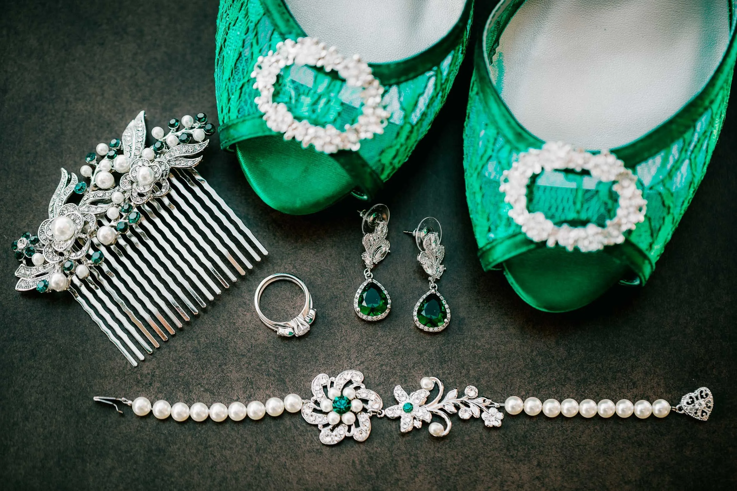 Green high-heeled shoes with clear buckle decoration, various jewelry pieces including pearl and emerald earrings, a rhinestone hair comb, pearl necklaces, and a silver ring, all arranged on a black surface.