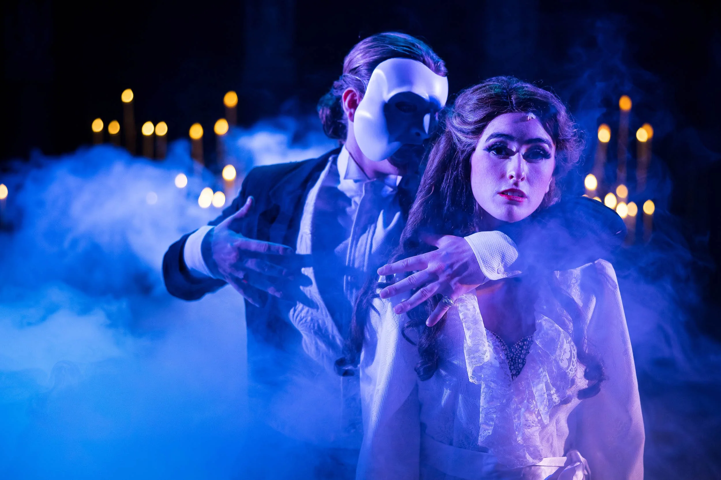 A man wearing a white mask and a woman with wavy hair in a white dress are depicted in a dark, smoky scene with blue and purple lighting, during a performance of Phantom of the Opera.