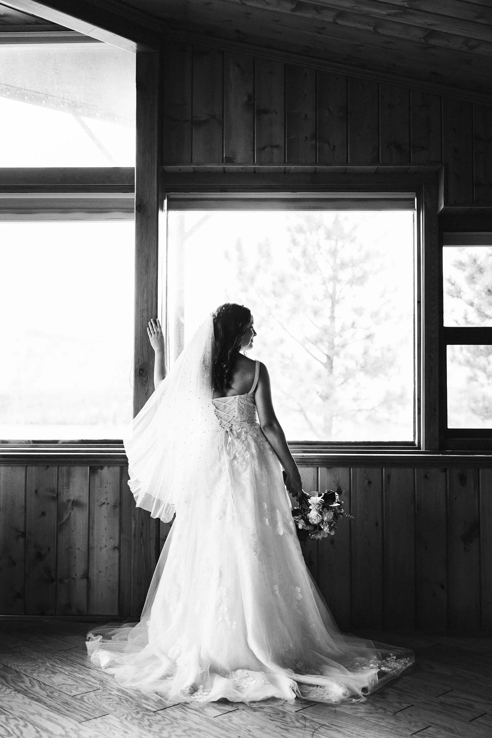 A bride in a wedding dress holding a bouquet stands in front of large windows, with wooden walls and floor, in a black-and-white photo.