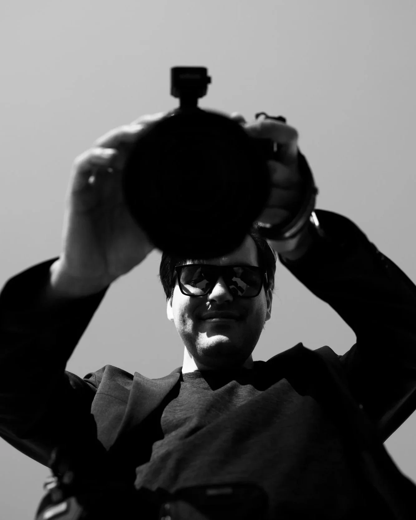 A person holding a camera pointed at the viewer, wearing sunglasses, against a plain sky background.
