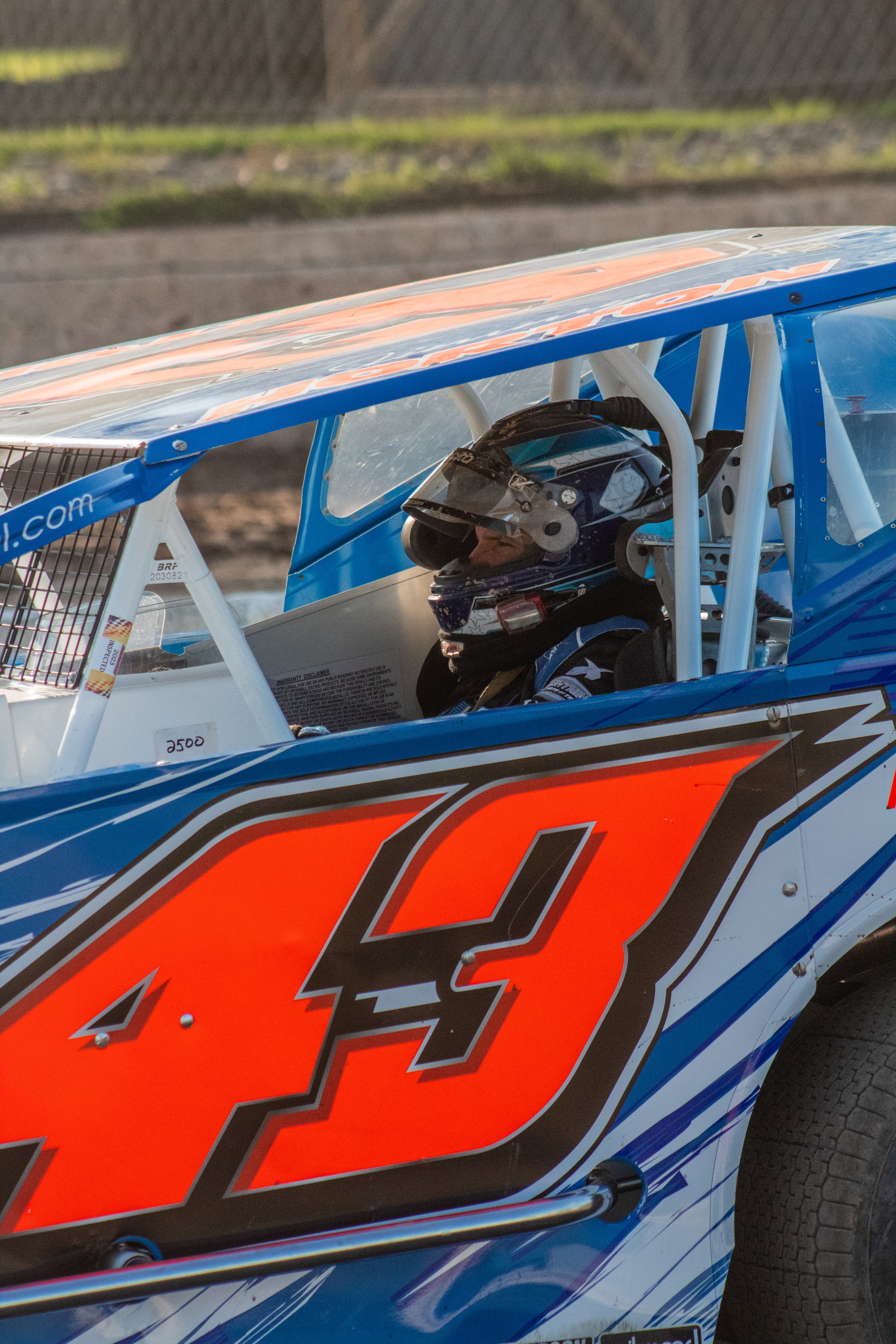 A race car with the number 43, blue and white paint, and a driver inside wearing a helmet, at a dirt race track.