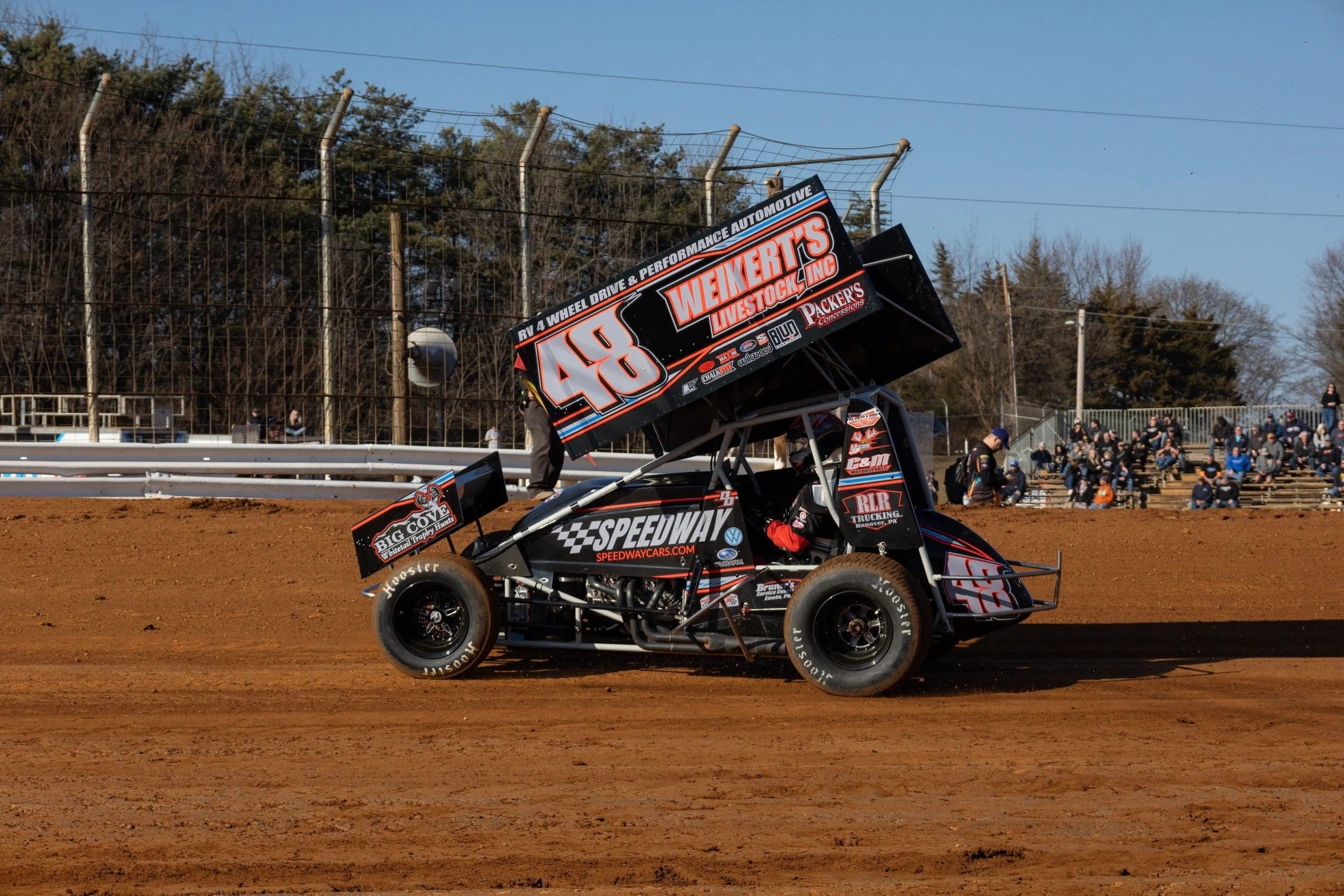 A sprint car racing on a dirt track with a driver inside, and spectators sitting on bleachers in the background.