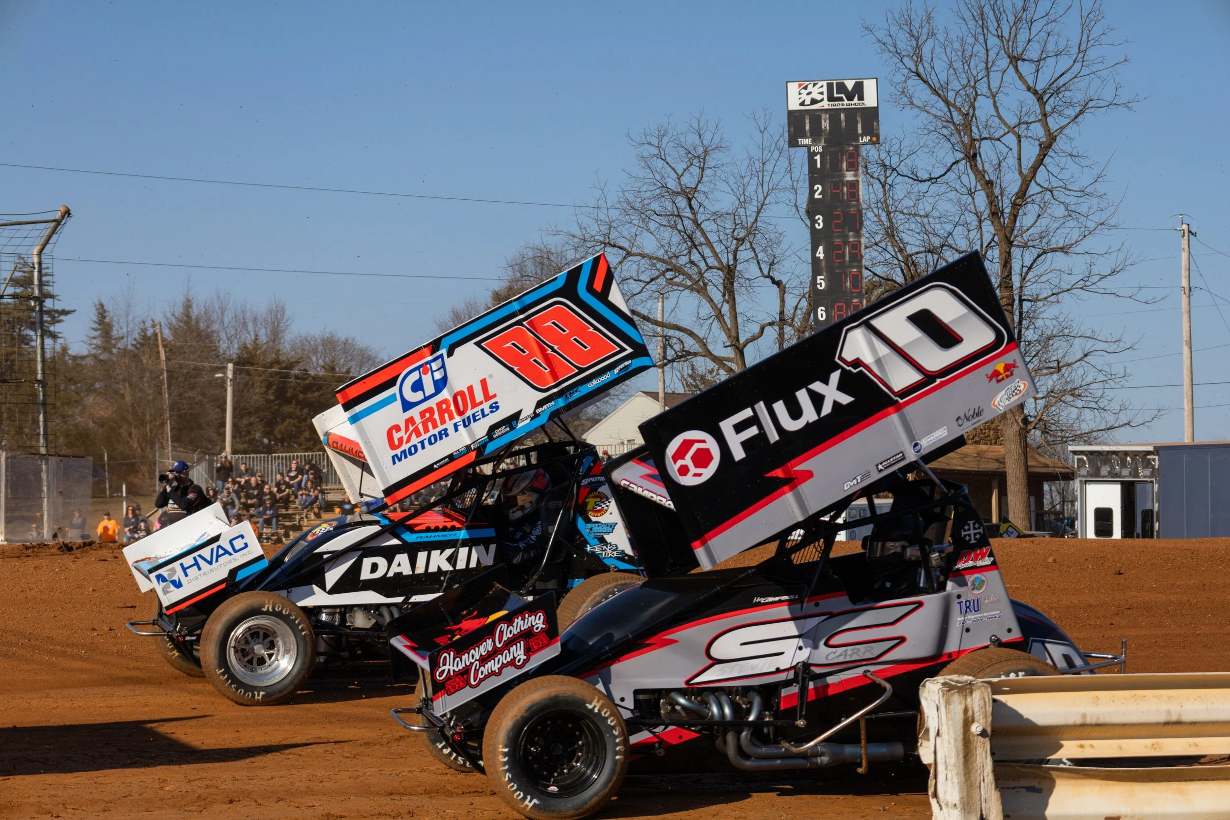 Two sprint cars racing on a dirt track during a race, with a scoreboard in the background displaying lap times and positions.