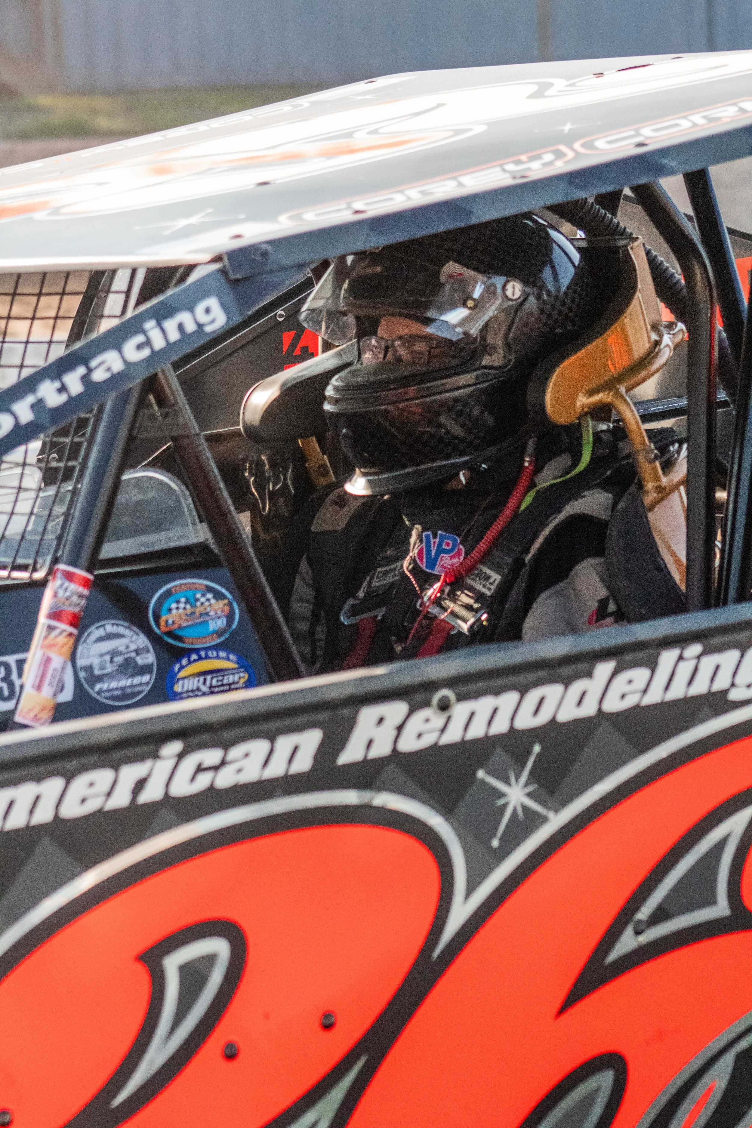 Race car driver sitting inside a race car with a black helmet and racing gear, visible logo stickers on the car door, including 'American Remodeling' and a large number '03' in red.