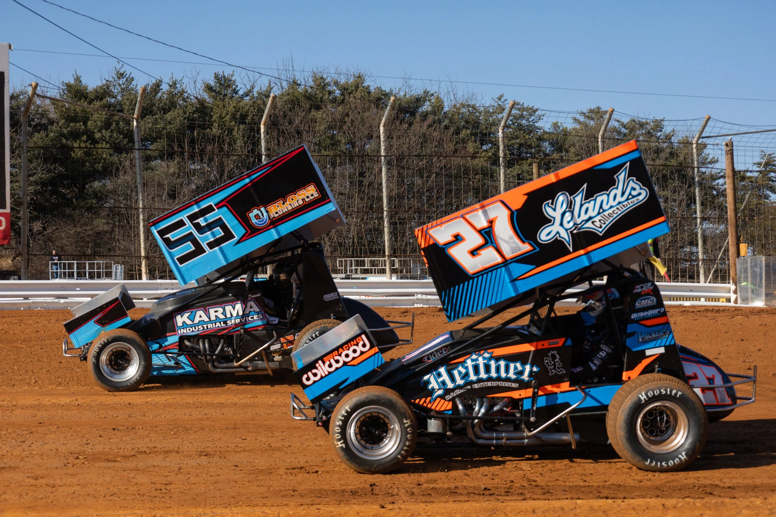Two sprint cars with large rear wings racing on a dirt track, with trees and a fence in the background.