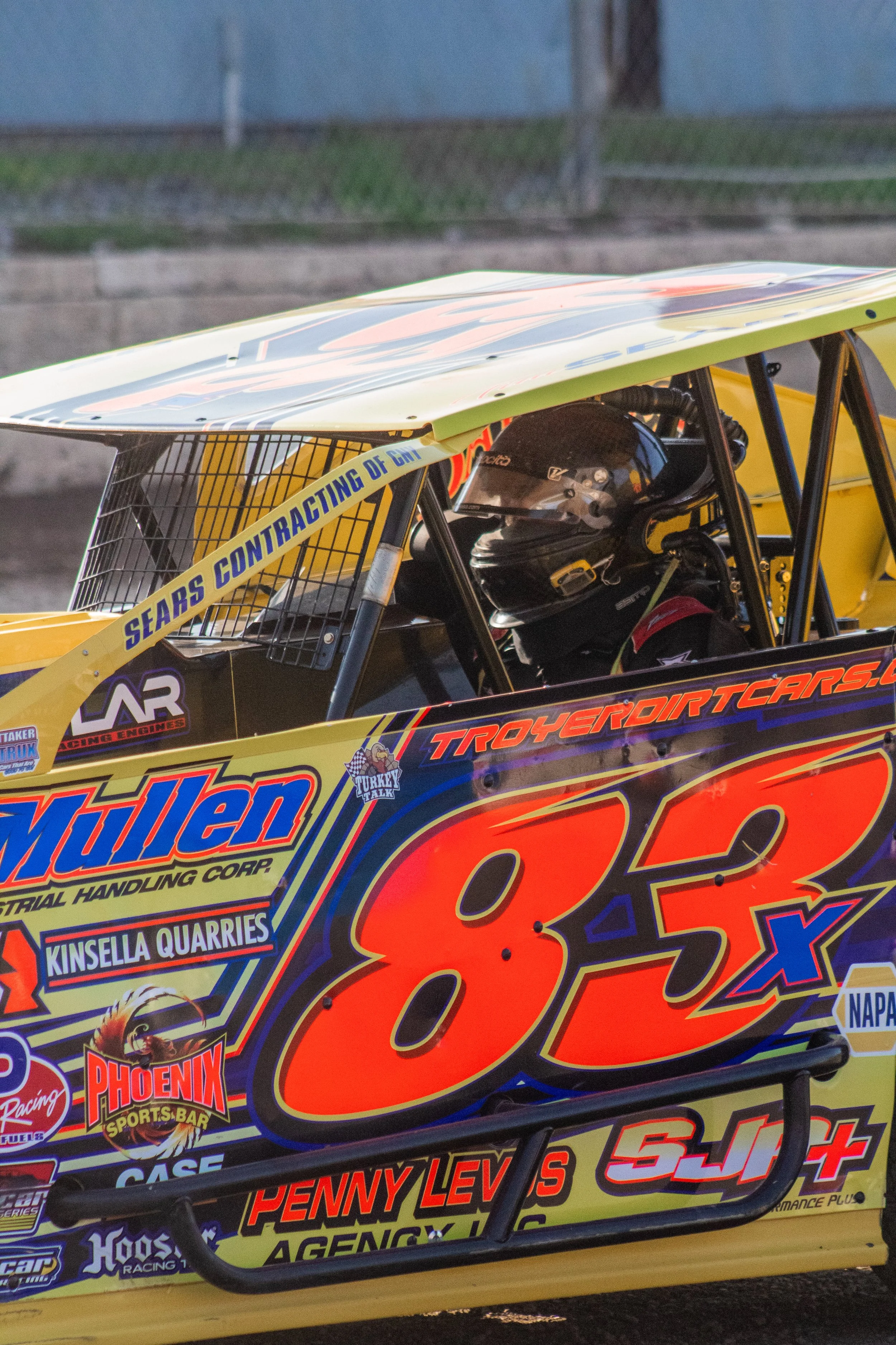 Close-up of a race car with vivid yellow and black colors, featuring the number 83 in large orange and black font. The car has various sponsor logos, including Miller, Penny Lewis Agency, and Phoenix Sports Bar, and a driver wearing a helmet seated i