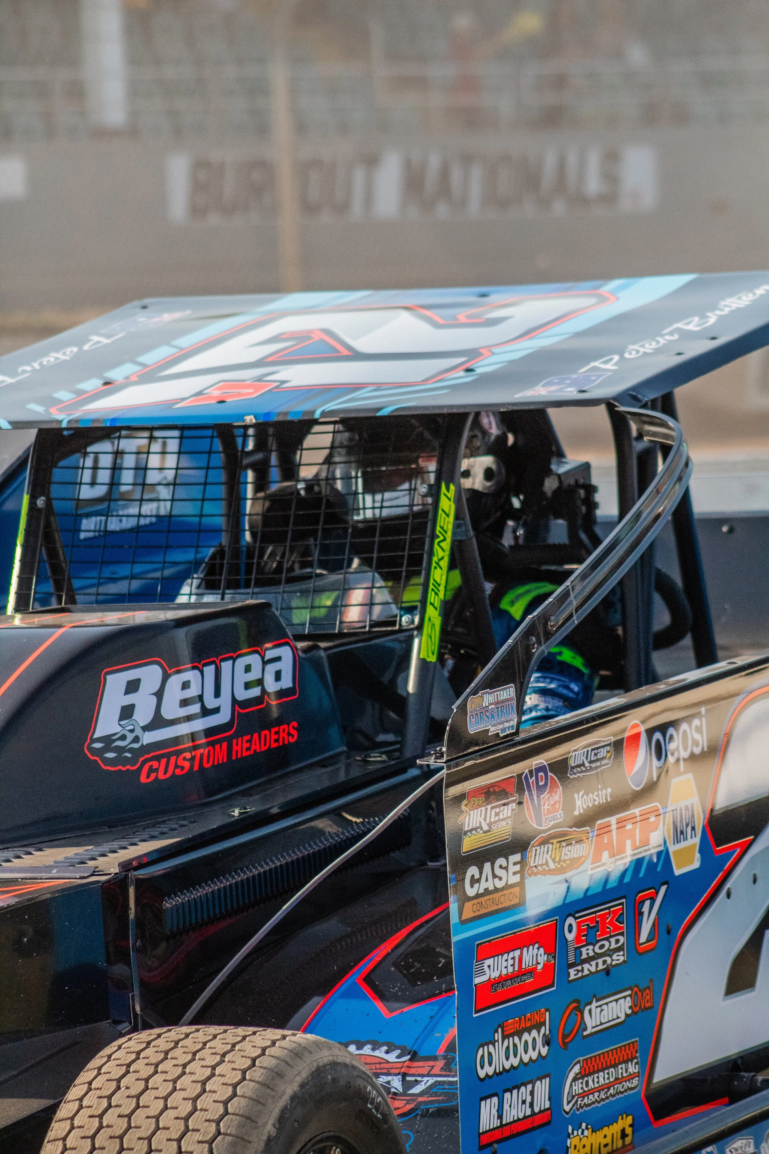 Close-up of a race car with sponsor logos, driver inside wearing helmet, on a dirt track.