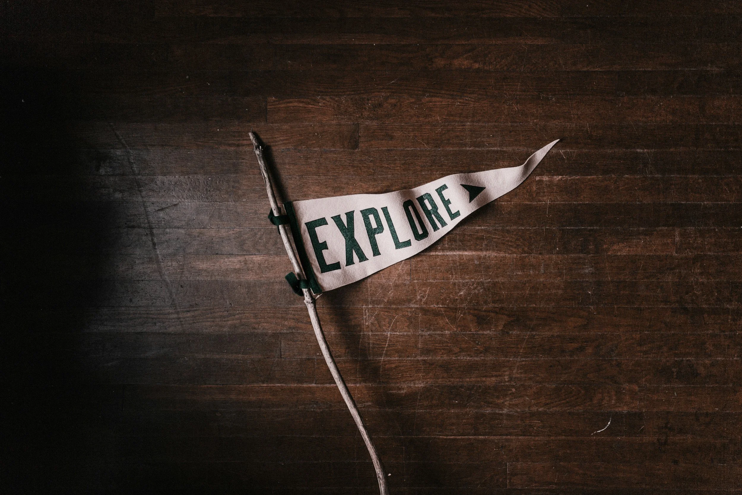 A small flag with the word "EXPLORE" on it, attached to a stick, resting on a wooden surface representing that clients are welcome to explore Lesley Keys coaching services and contact for more details