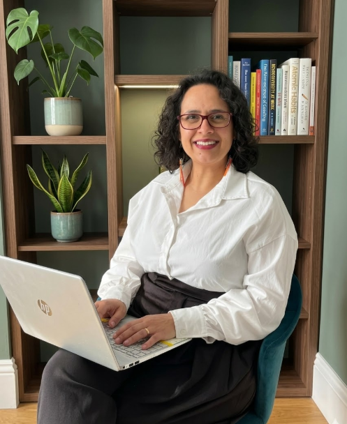 Lesley Keys Coach with curly dark hair and glasses smiling while sitting in a teal chair, working on a laptop in front of a bookshelf with potted plants and books.