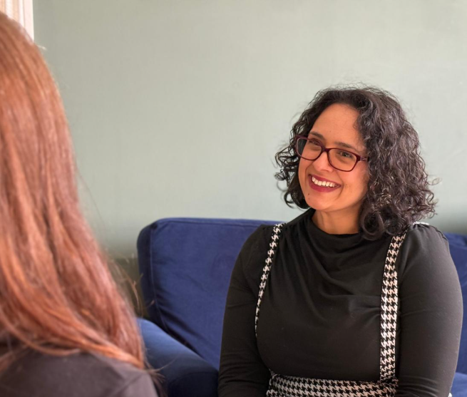 Lesley Keys Coach with curly dark hair, wearing glasses, smiling, open body language and listening encouragingly to a client with red hair in a room with light green walls and a blue couch.