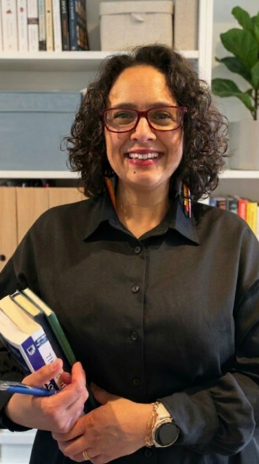 Lesley Keys Coach.  Dark curly hair, wearing glasses and a black shirt, holding a book in her left hand, standing in front of a bookshelf and a plant.
