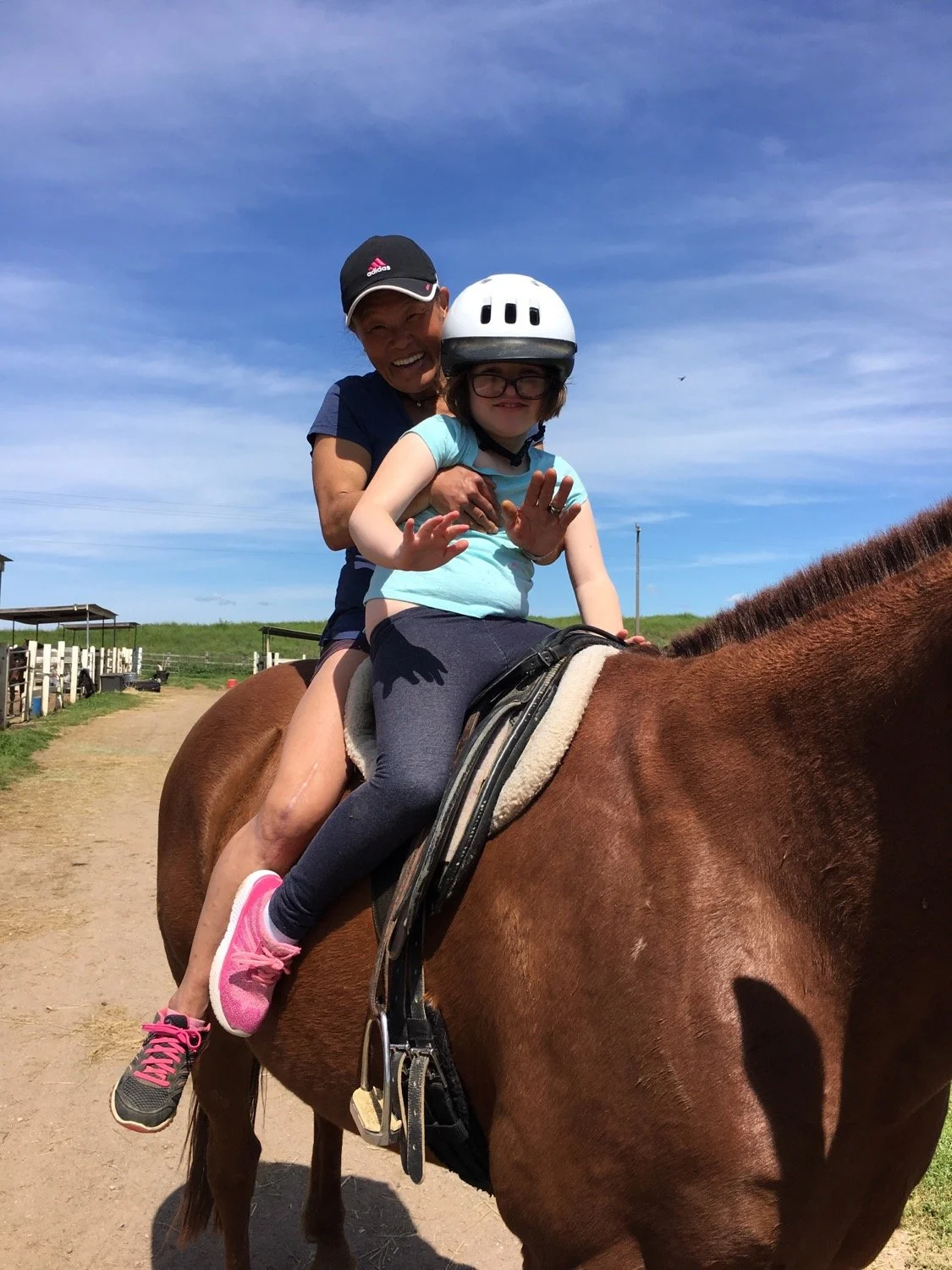 A woman and a girl riding a horse outdoors on a sunny day, with the woman sitting behind the girl, who is wearing a riding helmet and glasses, while both smile and wave.