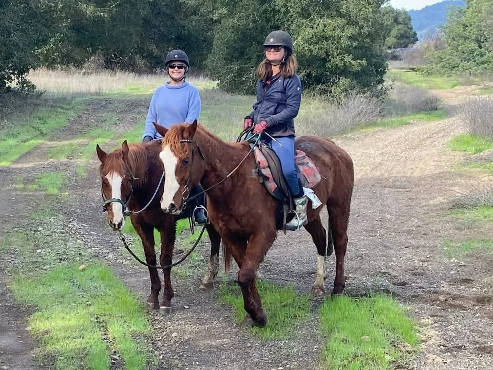 Two women riding horses on a dirt path in a rural area, surrounded by green grass and trees.