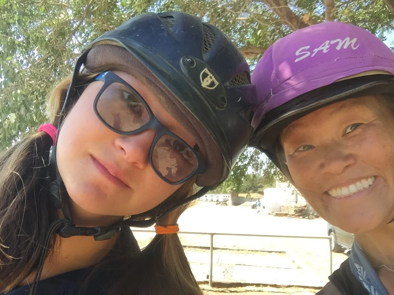 Two women wearing bike helmets, one with glasses and the other with a purple helmet, posing for a selfie outdoors with trees in the background.
