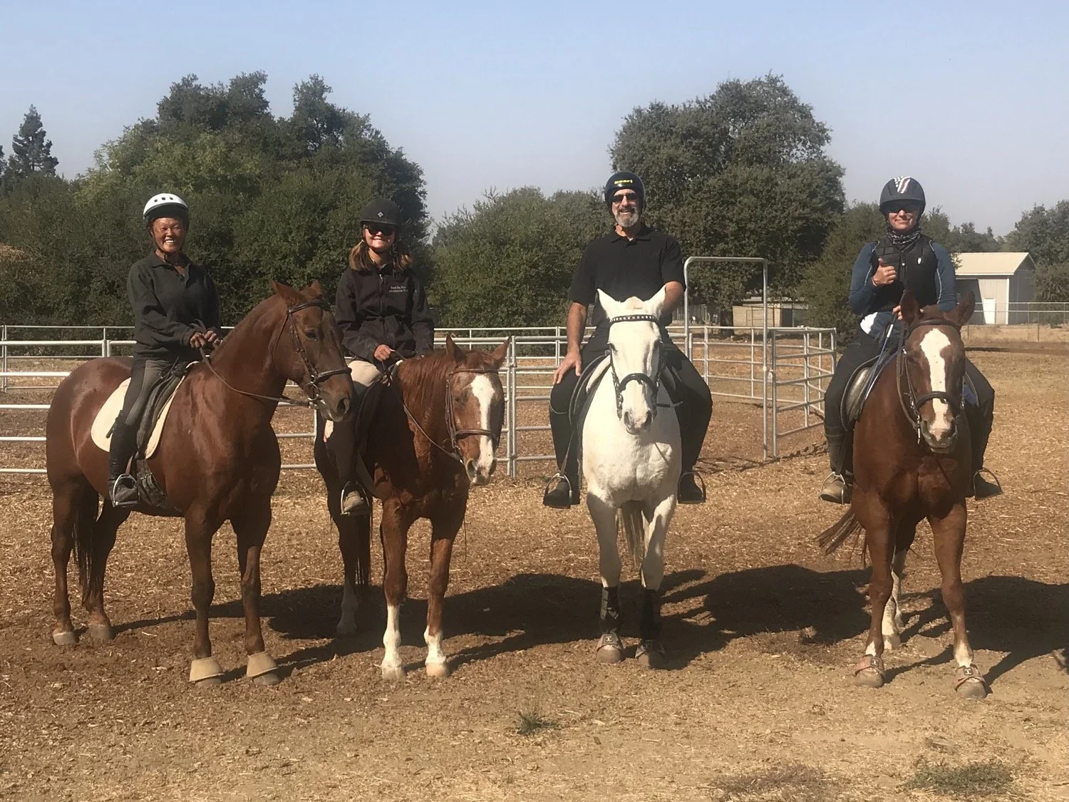 Four people riding horses in an outdoor horse riding arena, smiling and wearing safety helmets, with trees and a small building in the background.