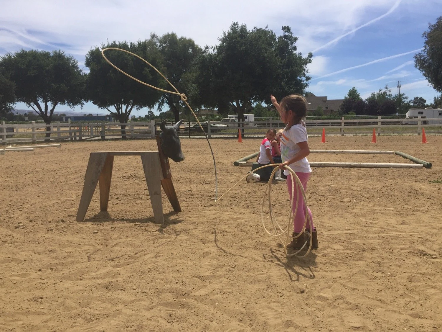 Girl with pink leggings and white t-shirt practicing roping with a wooden horse head target at an outdoor riding arena.