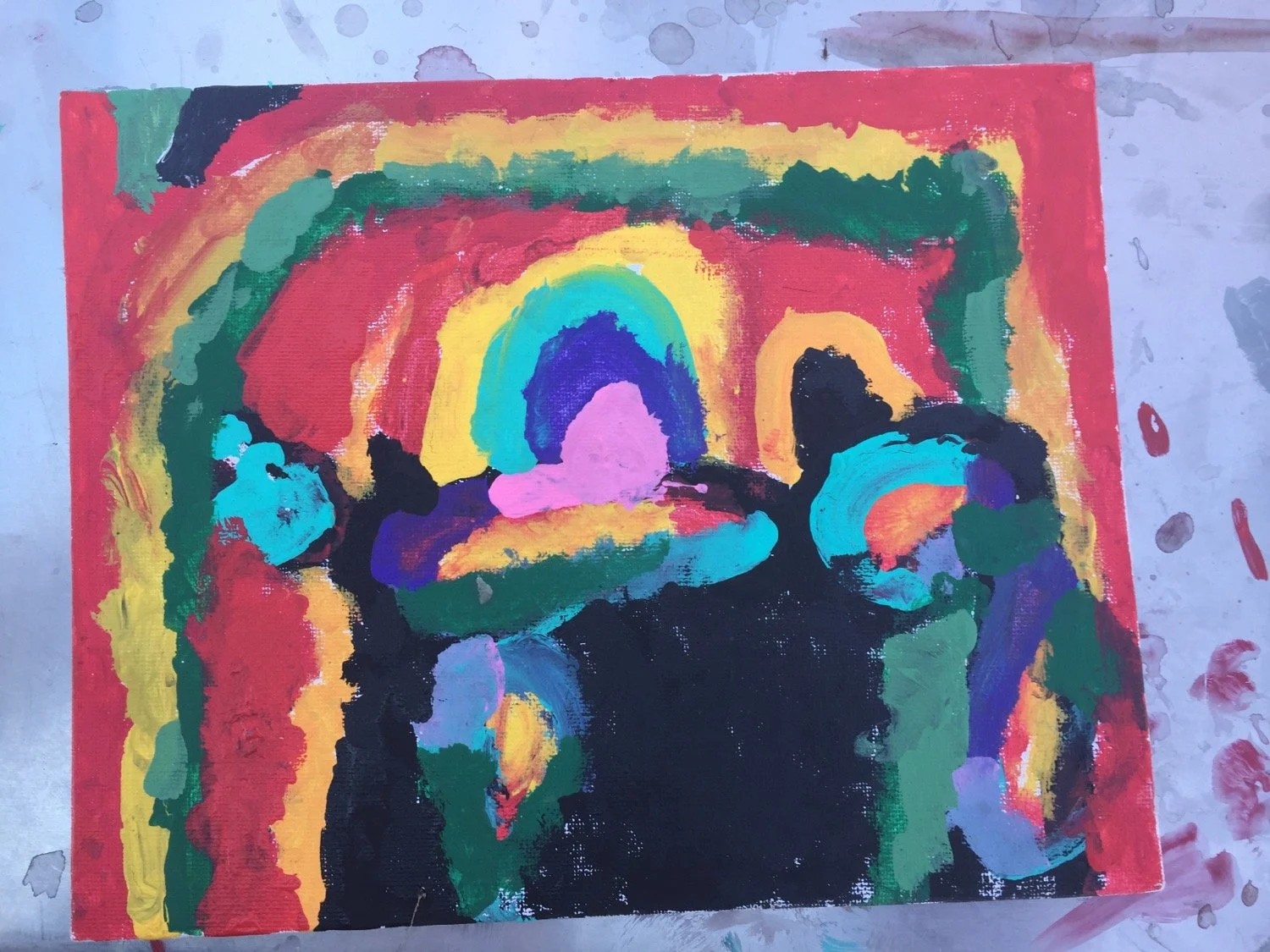 Colorful abstract painting with rainbow-like arches and figures, on a pink border, placed on a paint-spattered surface.