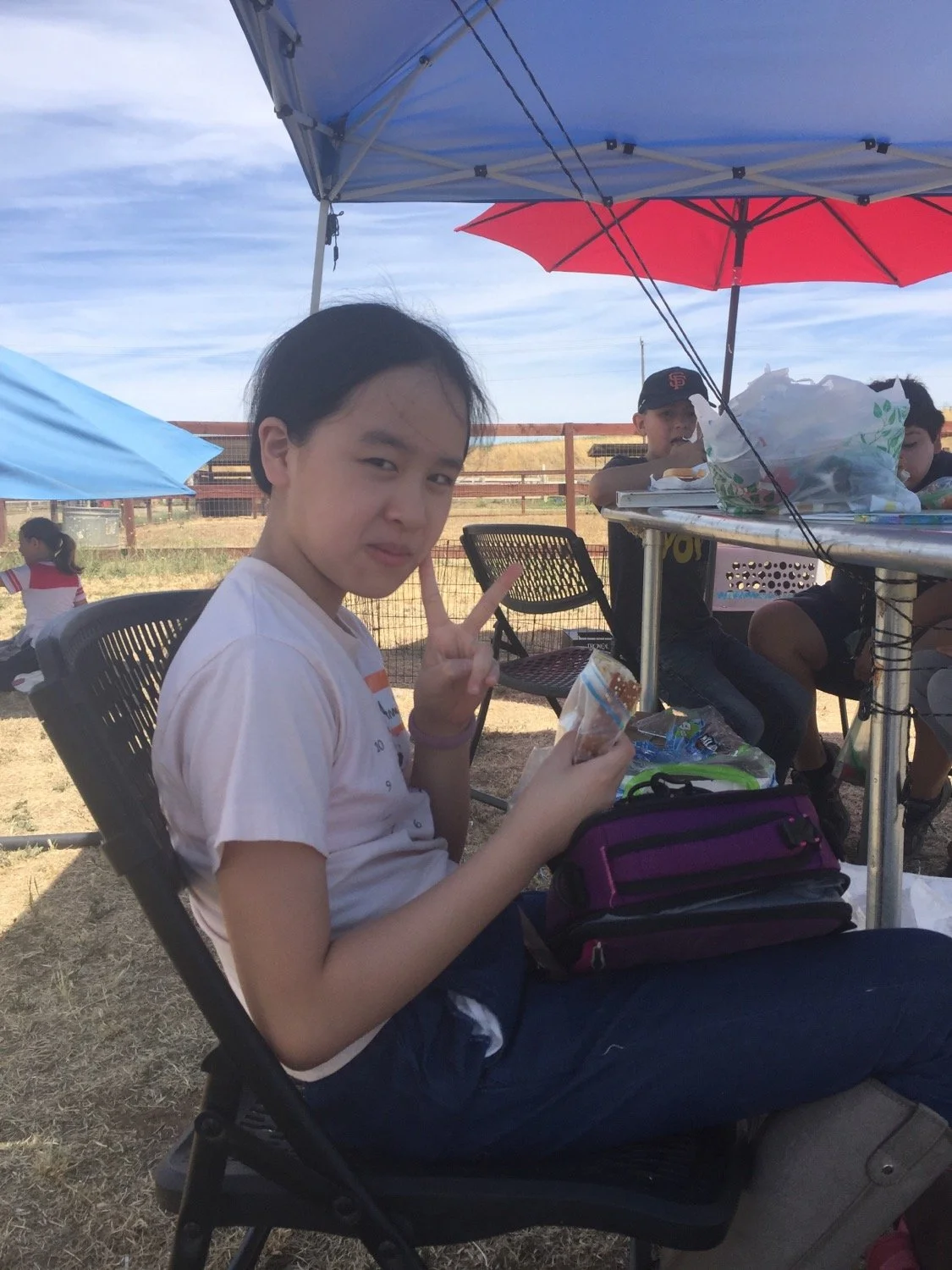 A young girl sitting in a folding chair under a blue canopy, making a peace sign with her fingers, holding a snack, with other children and umbrellas in the background outdoors on a sunny day.