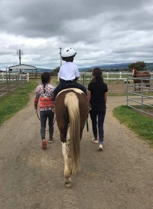 Child wearing a helmet riding a pony, accompanied by two women walking alongside on a farm trail under cloudy skies.
