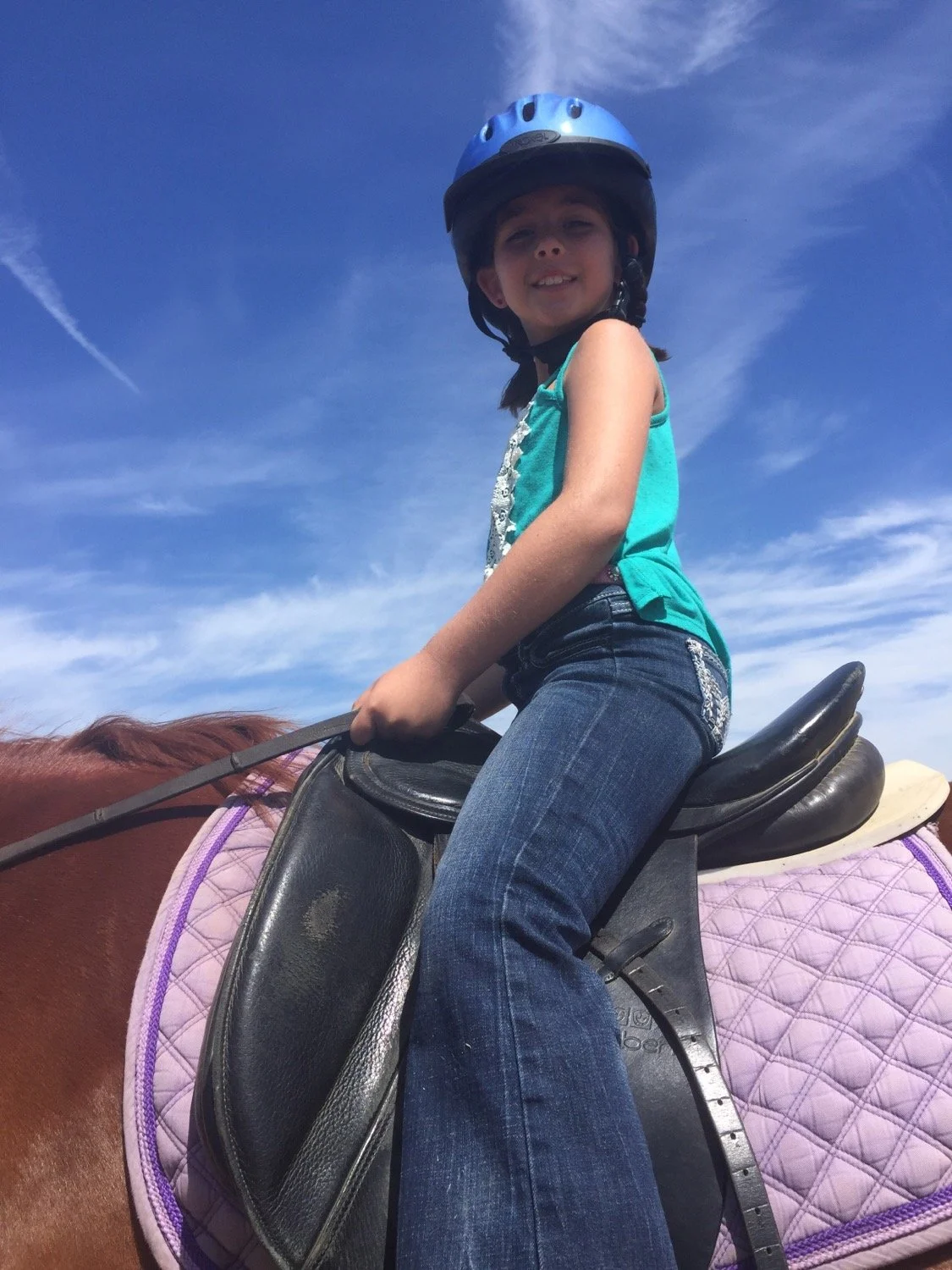 Child girl riding a horse with a helmet, sitting on a saddle, blue sky in the background.