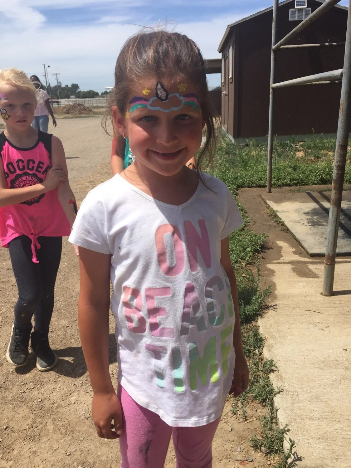 Smiling young girl with face paint standing outdoors on a sunny day, wearing a white T-shirt with colorful text and pink pants, with children and a building in the background.