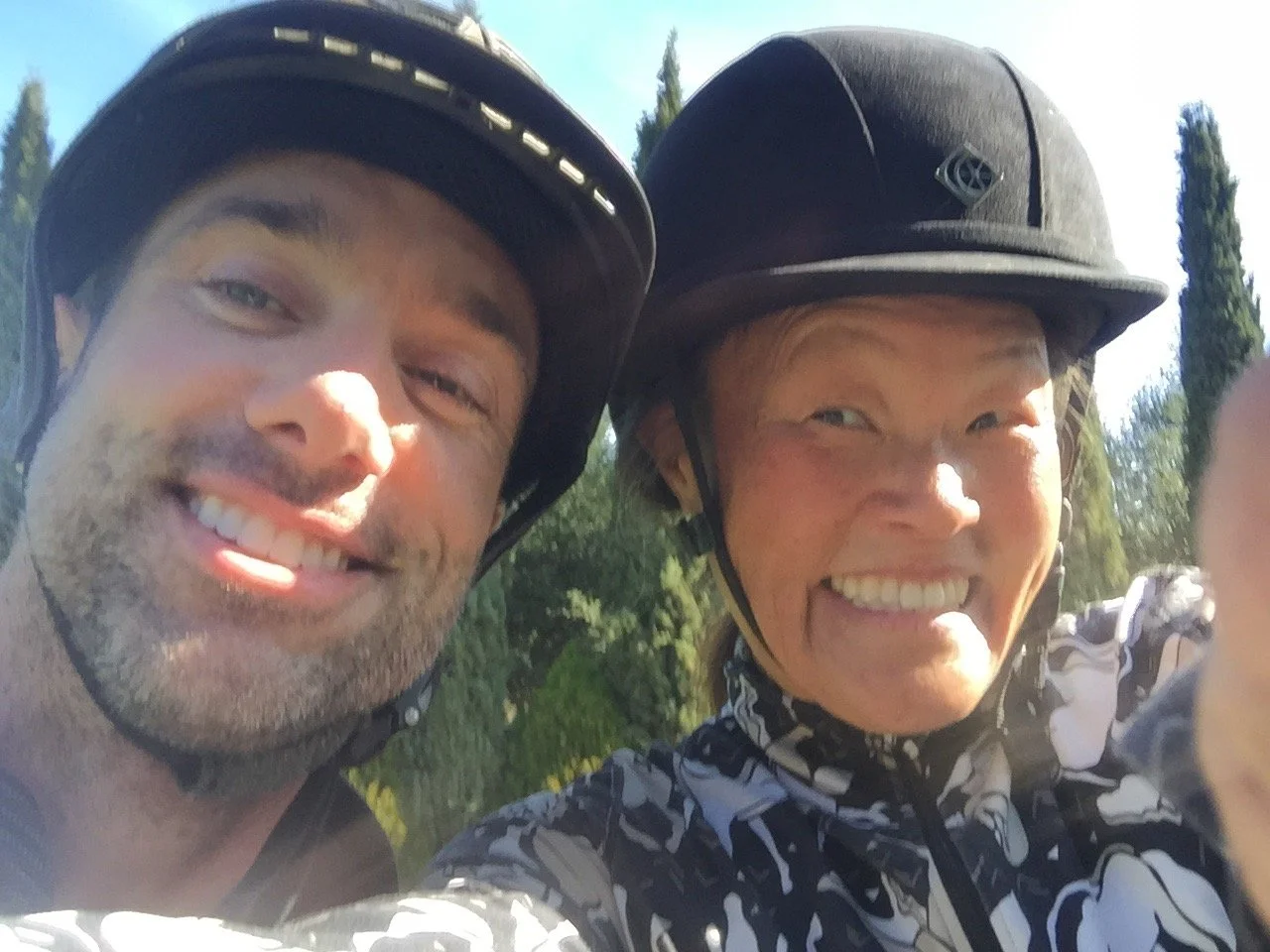 Two people smiling in outdoor setting, both wearing black helmets, with green trees in the background.