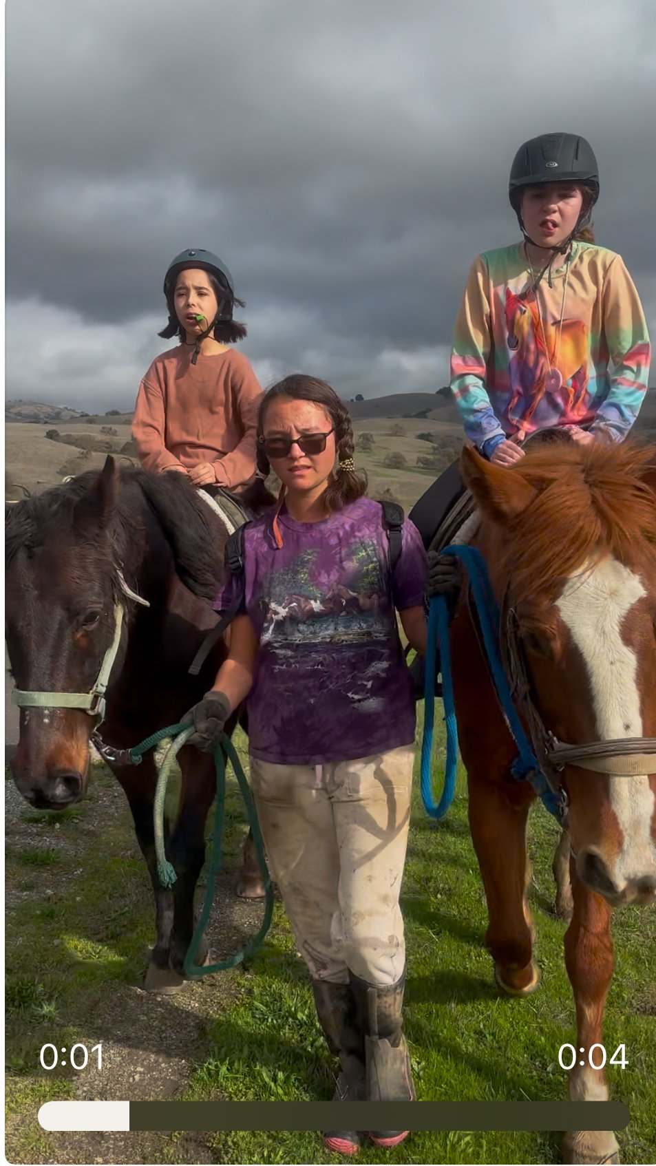 Three young girls riding horses outdoors on cloudy day, with grassy hills in the background.