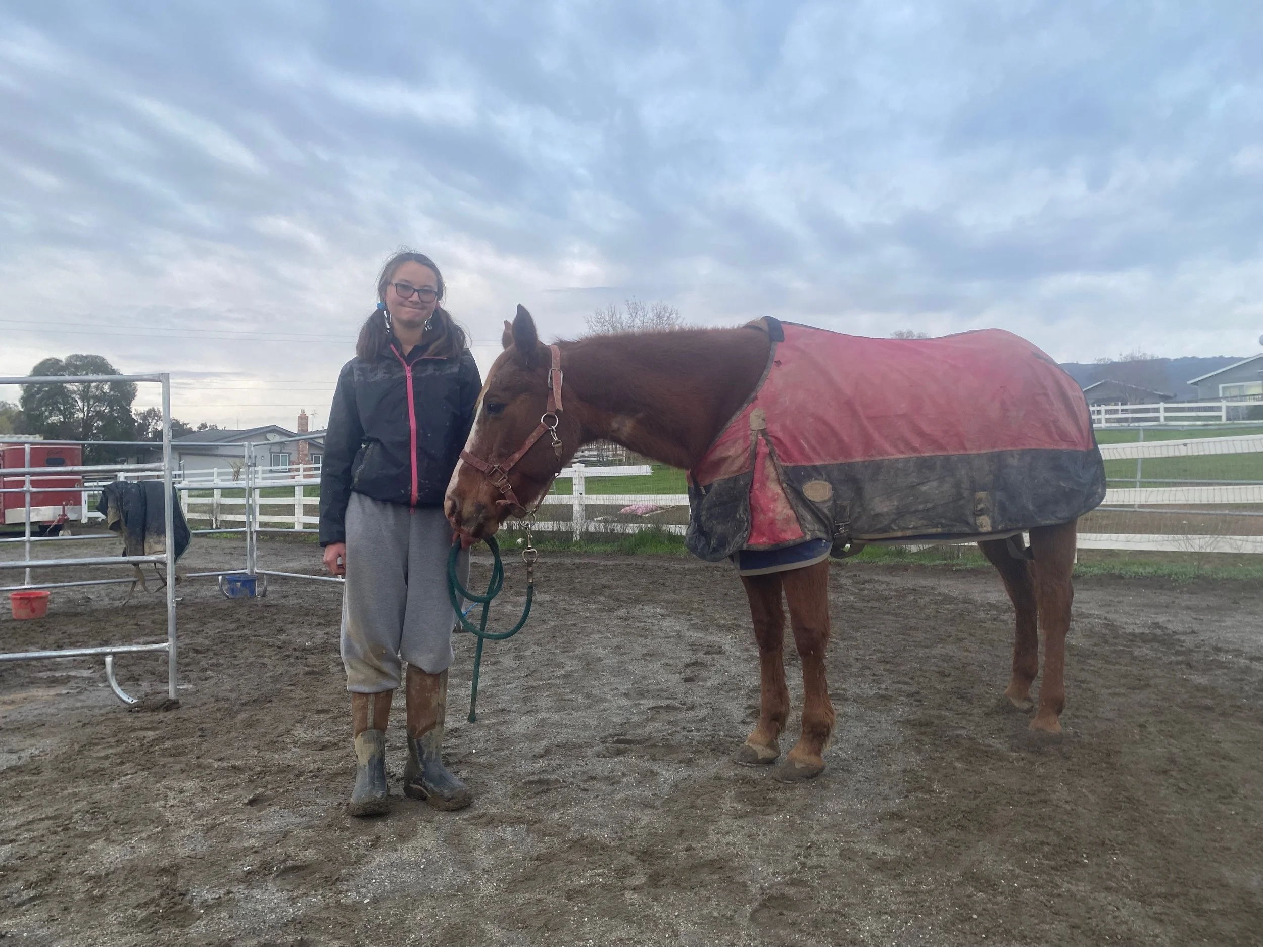 A girl in rain boots and a jacket standing next to a brown horse wearing a red and black blanket, in an outdoor paddock with a cloudy sky.