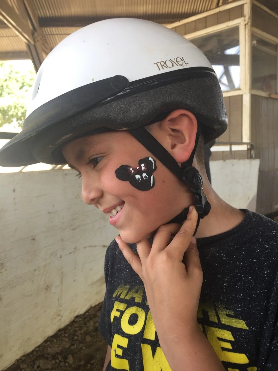A young boy wearing a white bike helmet with black padding. He has a painted face with a black mouse face, large ears, and white eyes. The boy is smiling and holding the chin strap of his helmet. He is wearing a dark gray t-shirt with yellow and whit