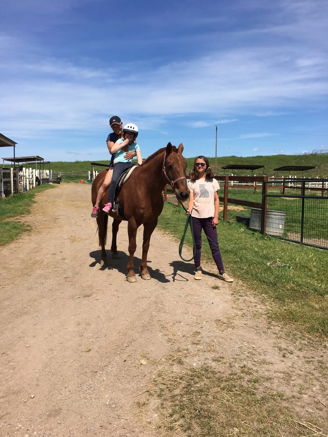 Two children riding a brown horse at a farm, with an adult woman standing nearby holding the horse's lead, under a clear blue sky.