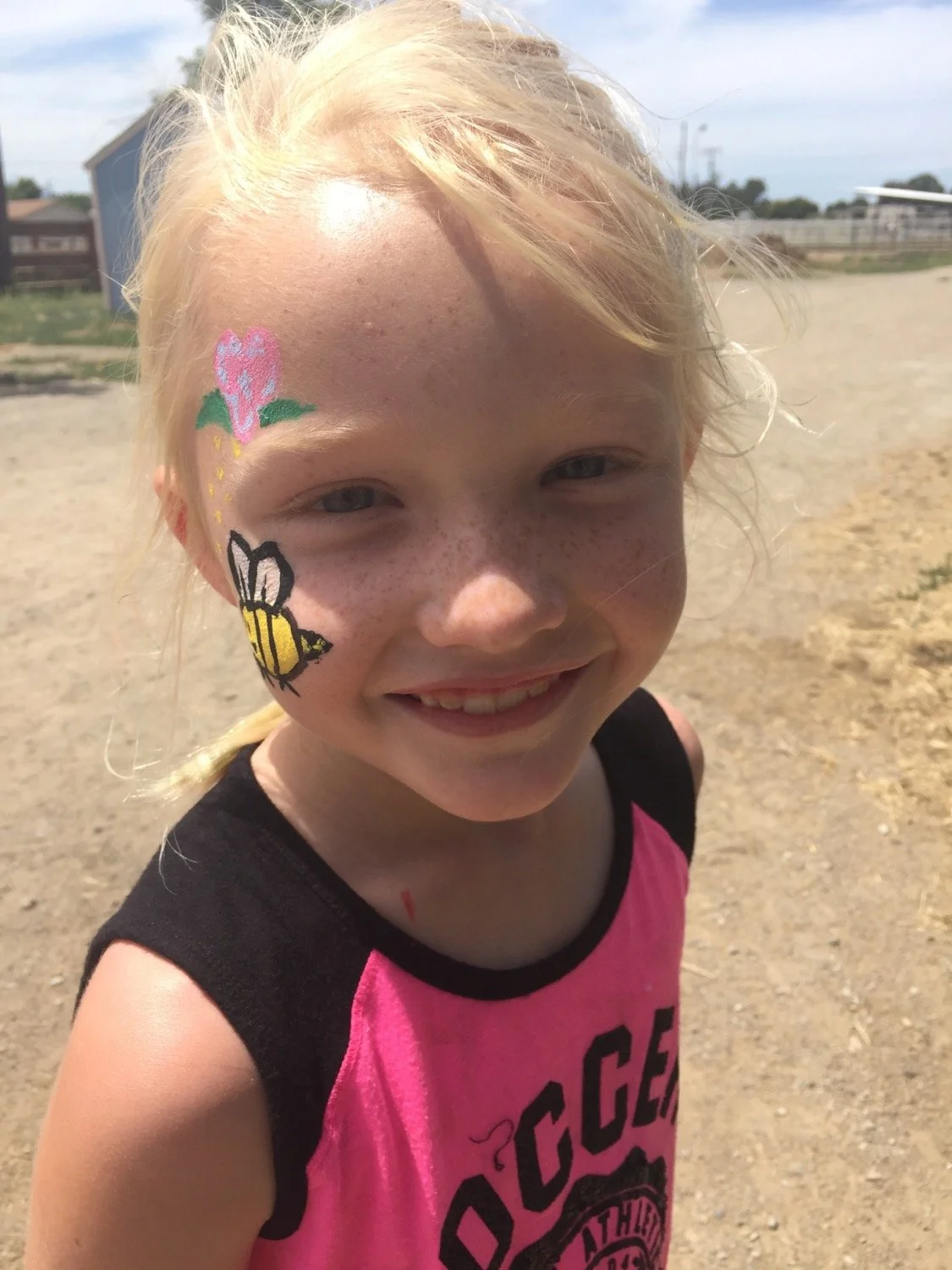 A young girl with blonde hair and freckles smiling outdoors. She has face paint of a pink flower and a yellow and black bee on her cheek. She is wearing a pink and black sleeveless shirt and standing on a dirt surface with a blue building and a fence