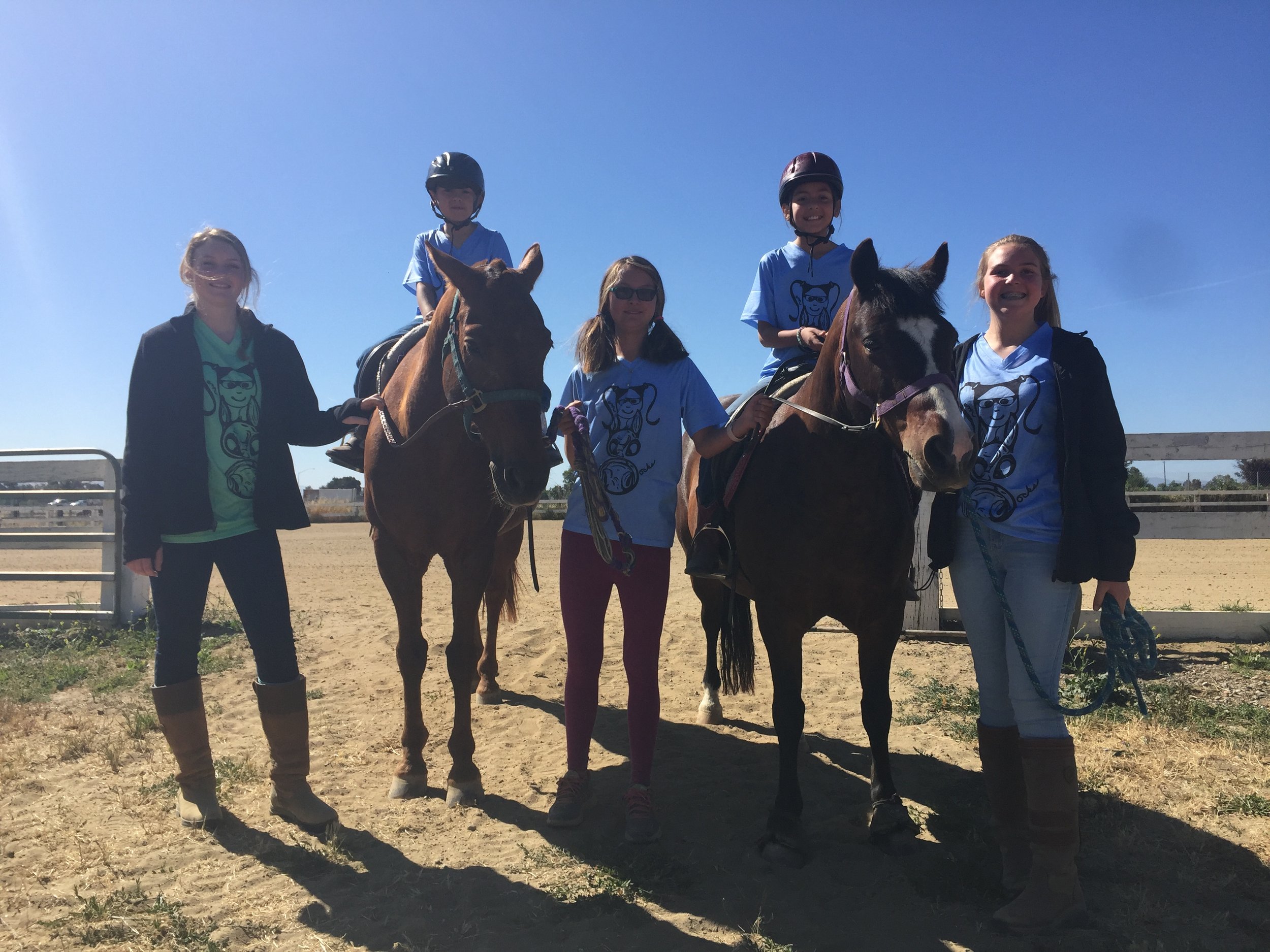 Five girls, three standing and two riding horses, posing outdoors in a dirt arena under a clear blue sky.