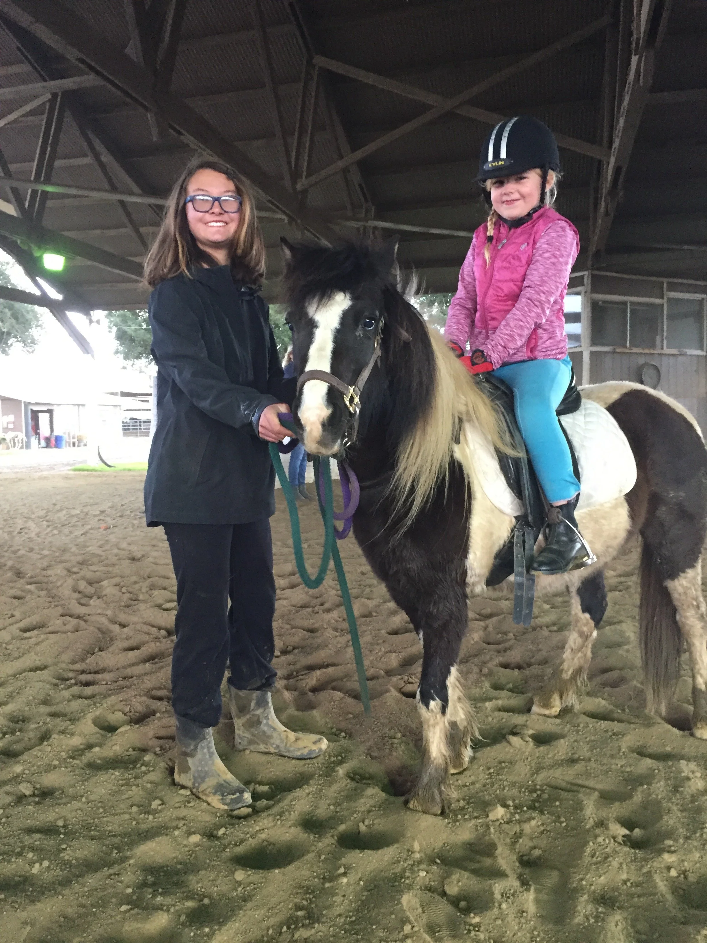A young girl riding a horse indoors, smiling, while an older girl holding the horse's bridle stands beside them, both girls smiling and wearing riding gear.