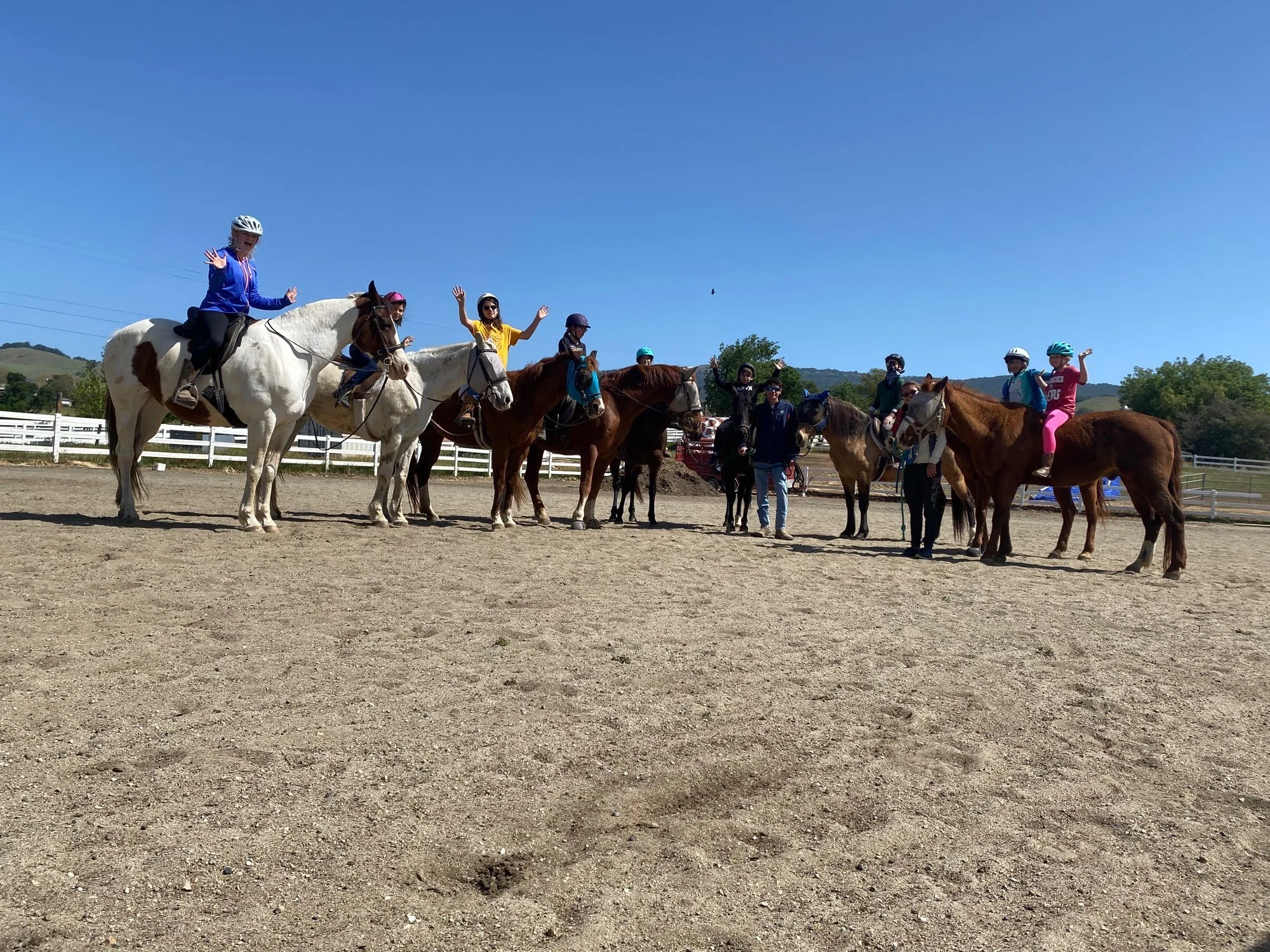 Group of children and adults on horseback, some waving, in an outdoor riding arena on a sunny day.