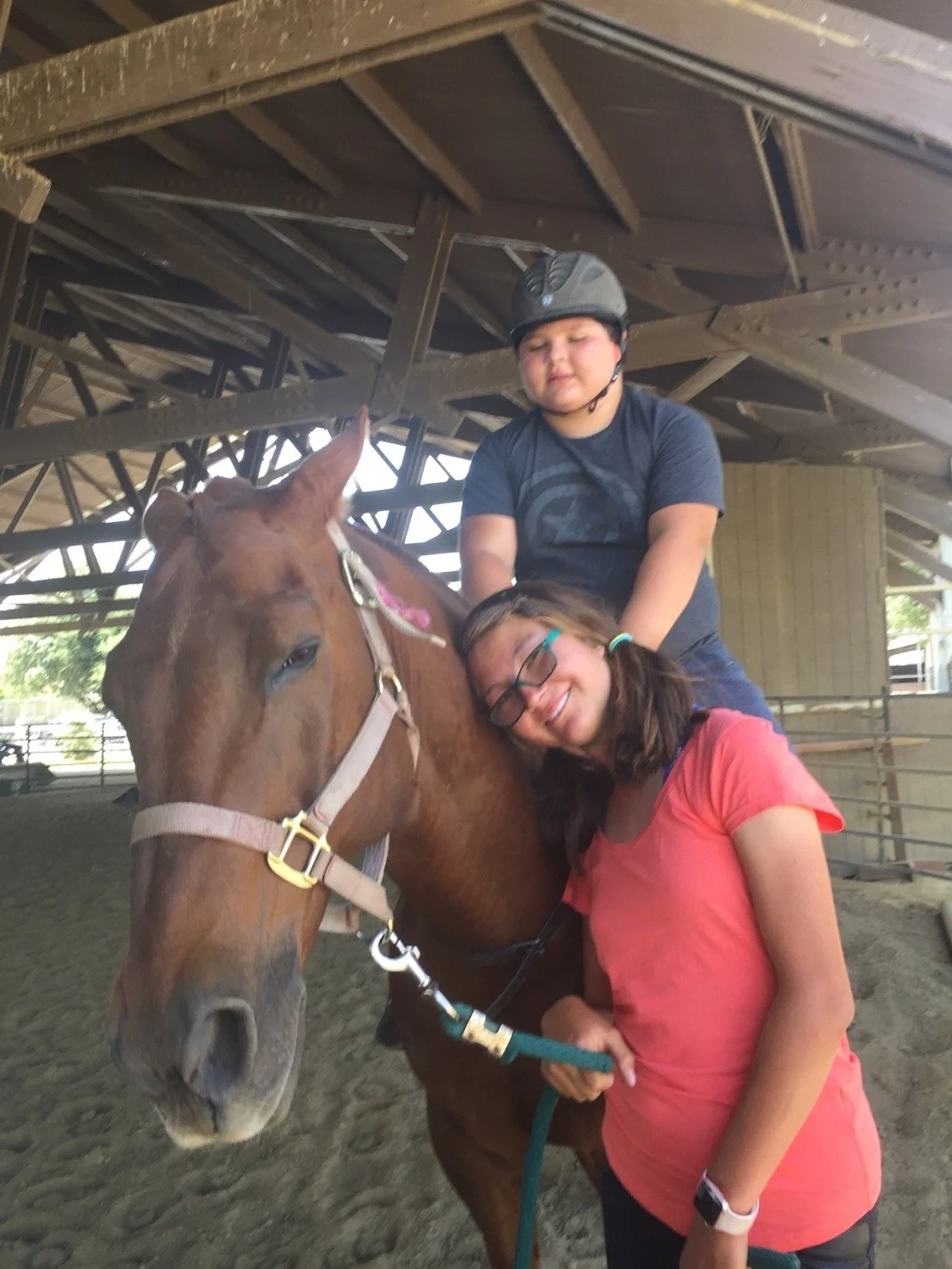A girl with glasses and a boy with a helmet holding a brown horse under a wooden shelter.