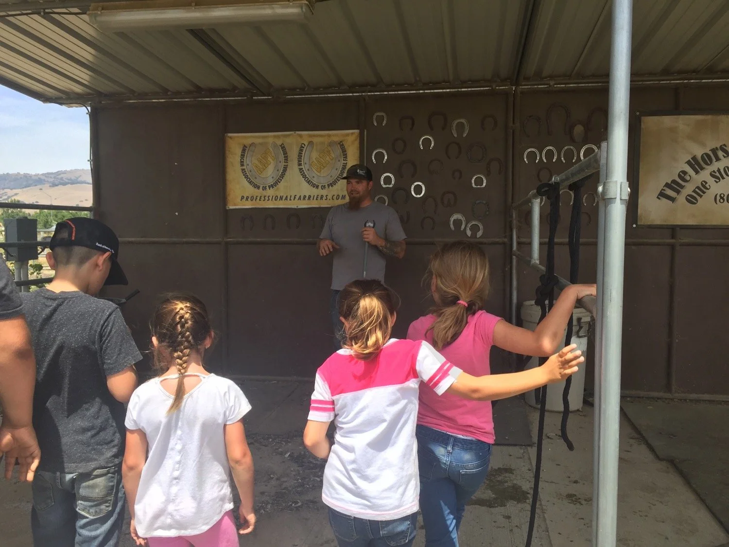 Children and a man inside a barn-like structure, with the children listening to a man with a microphone, suggesting a demonstration or presentation about animal handling or training.