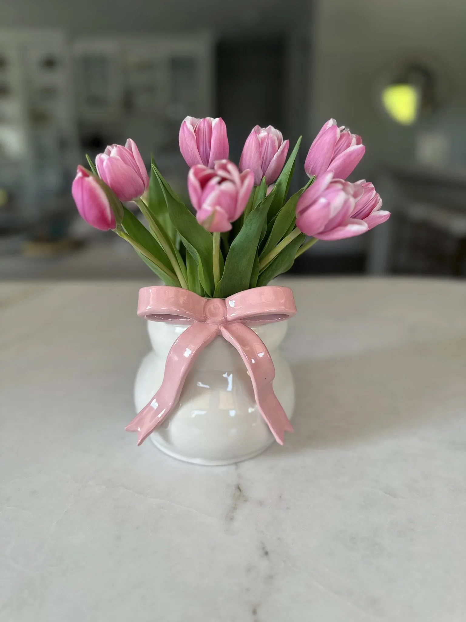 A small white vase with a pink ribbon tied around its neck, holding a bouquet of pink tulips with green leaves, placed on a light-colored surface.