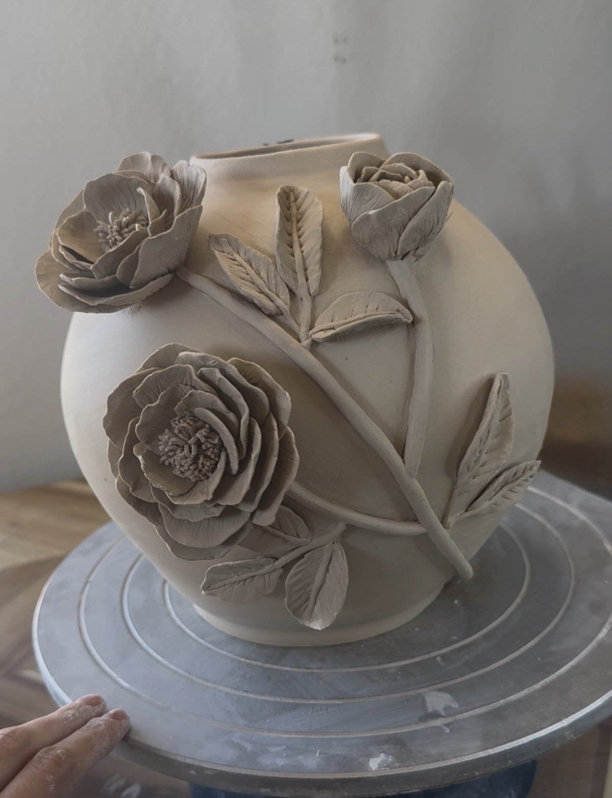 A white ceramic vase with intricate carved roses and leaves, placed on a turntable, with a person's hand in the lower left corner.