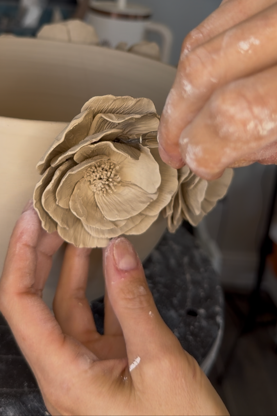 Close-up of hands shaping a beige, detailed clay flower sculpture in an art studio.