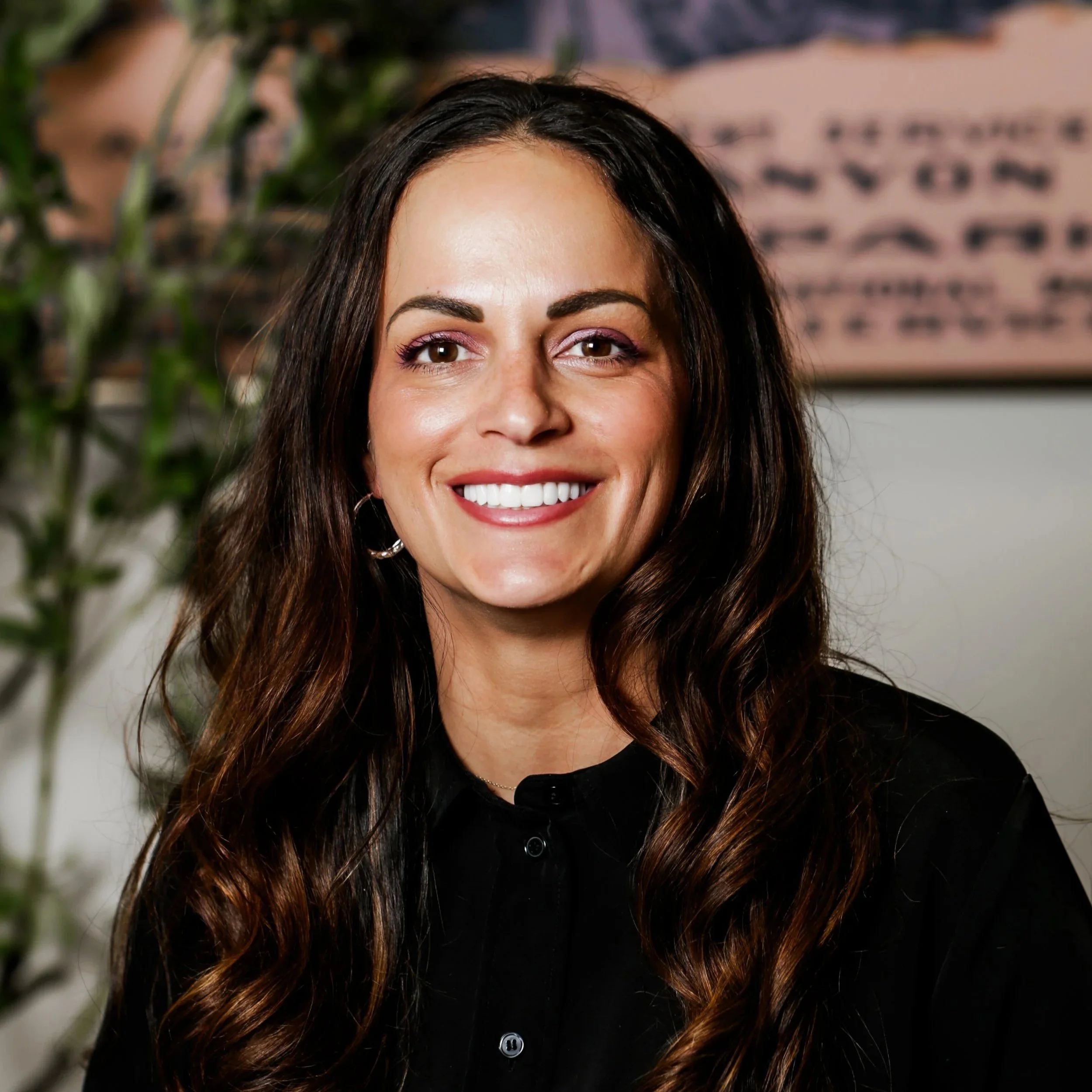 A smiling woman with long wavy brown hair, wearing a black top, standing indoors with a blurred background including a framed picture and a plant.