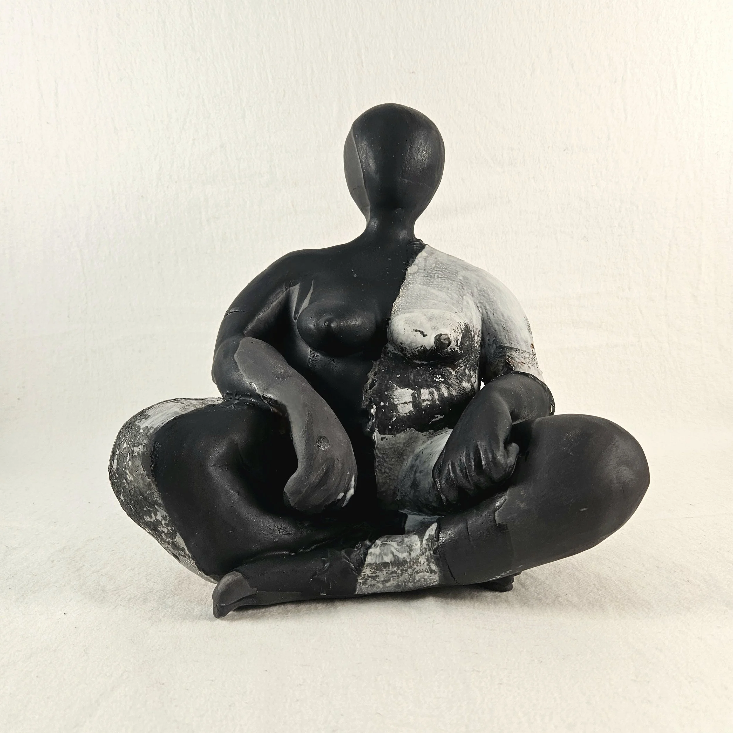Abstract black and white sculpture of a seated woman with crossed legs, with a split color contrast on the body, facing forward against a plain background.